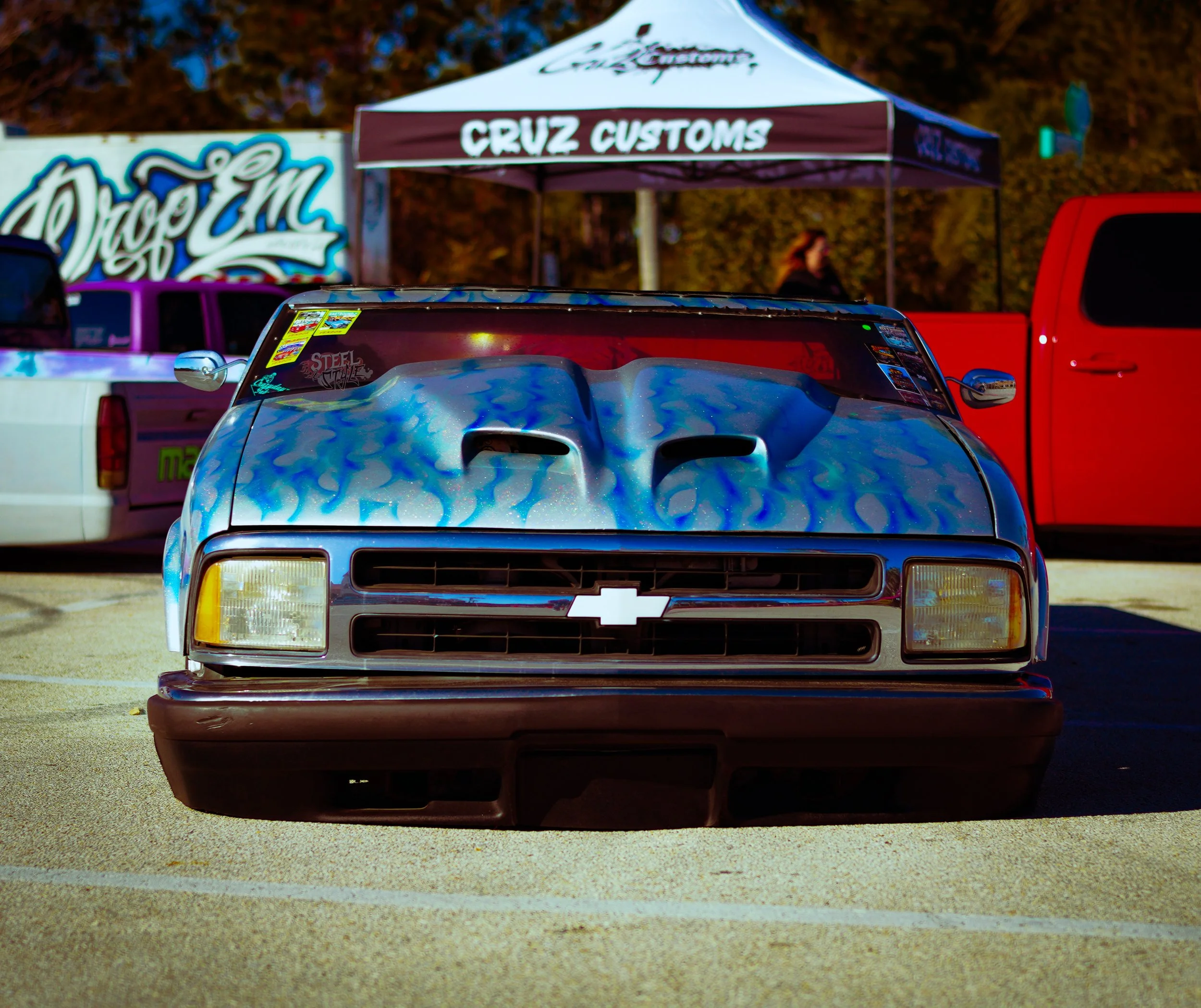 A Chevrolet race car with a blue flame paint design parked in a lot, with a Cruz Customs tent and other vehicles in the background.