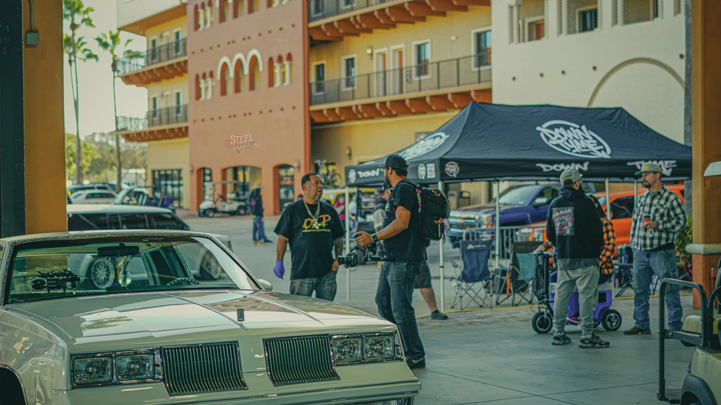 A person with a camera and glove talking to another person near a vintage car at an outdoor event with tents and people in the background.