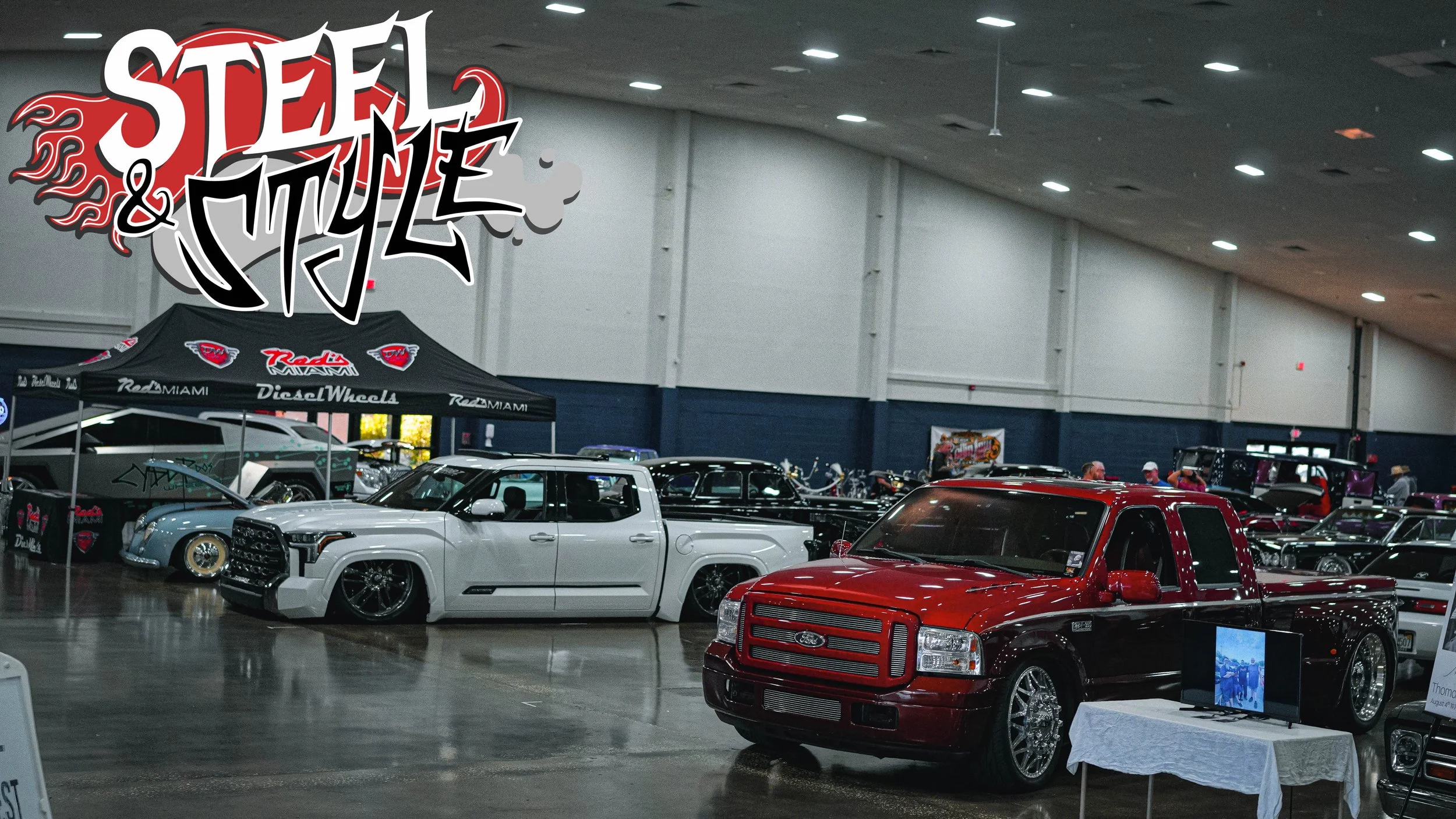 At an indoor car show, a large sign reads 'Stee & Style'. Several customized cars, including a white pickup truck and a red and black pickup truck, are on display. Tents and informational displays are set up around the space, with people walking and 