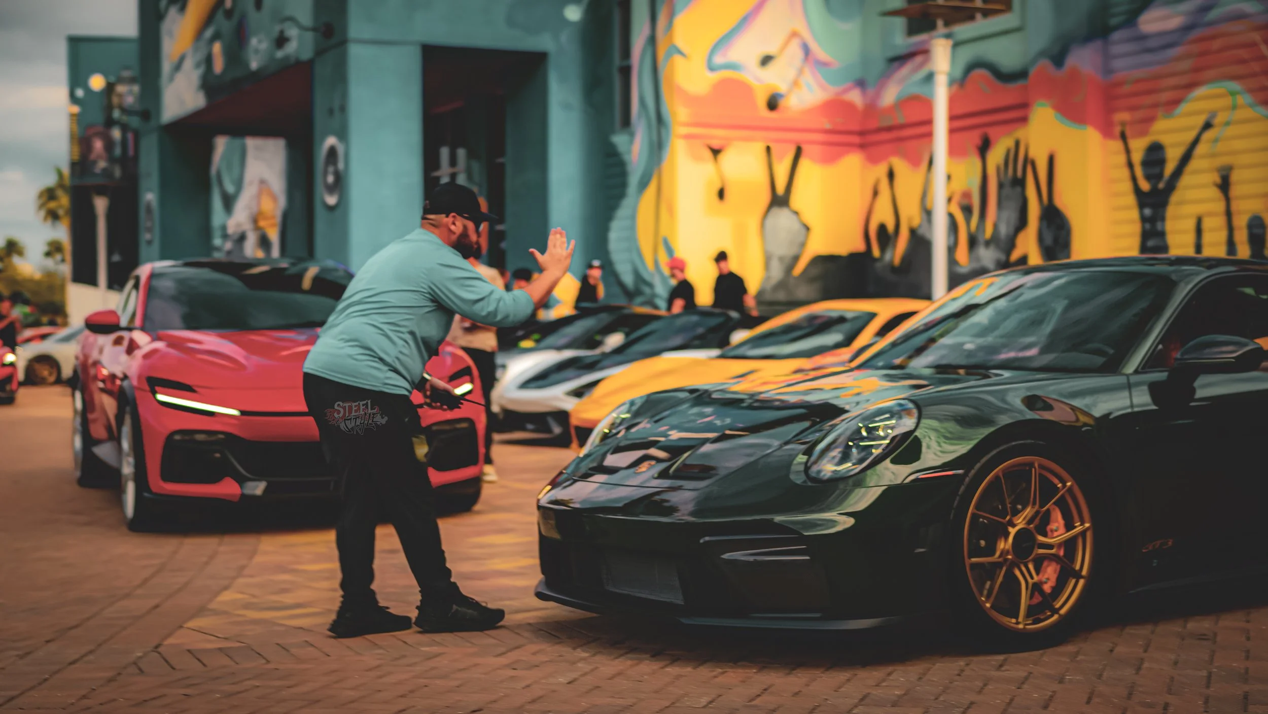 Person giving a high-five to a black Porsche sports car parked in front of a colorful graffiti mural at a car show.