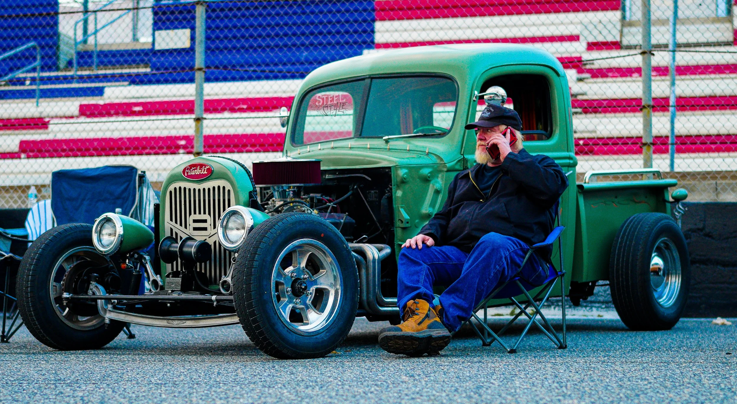 A man with a beard sitting in a blue folding chair next to a vintage green pickup truck, talking on a cell phone, with a racetrack grandstand in the background.