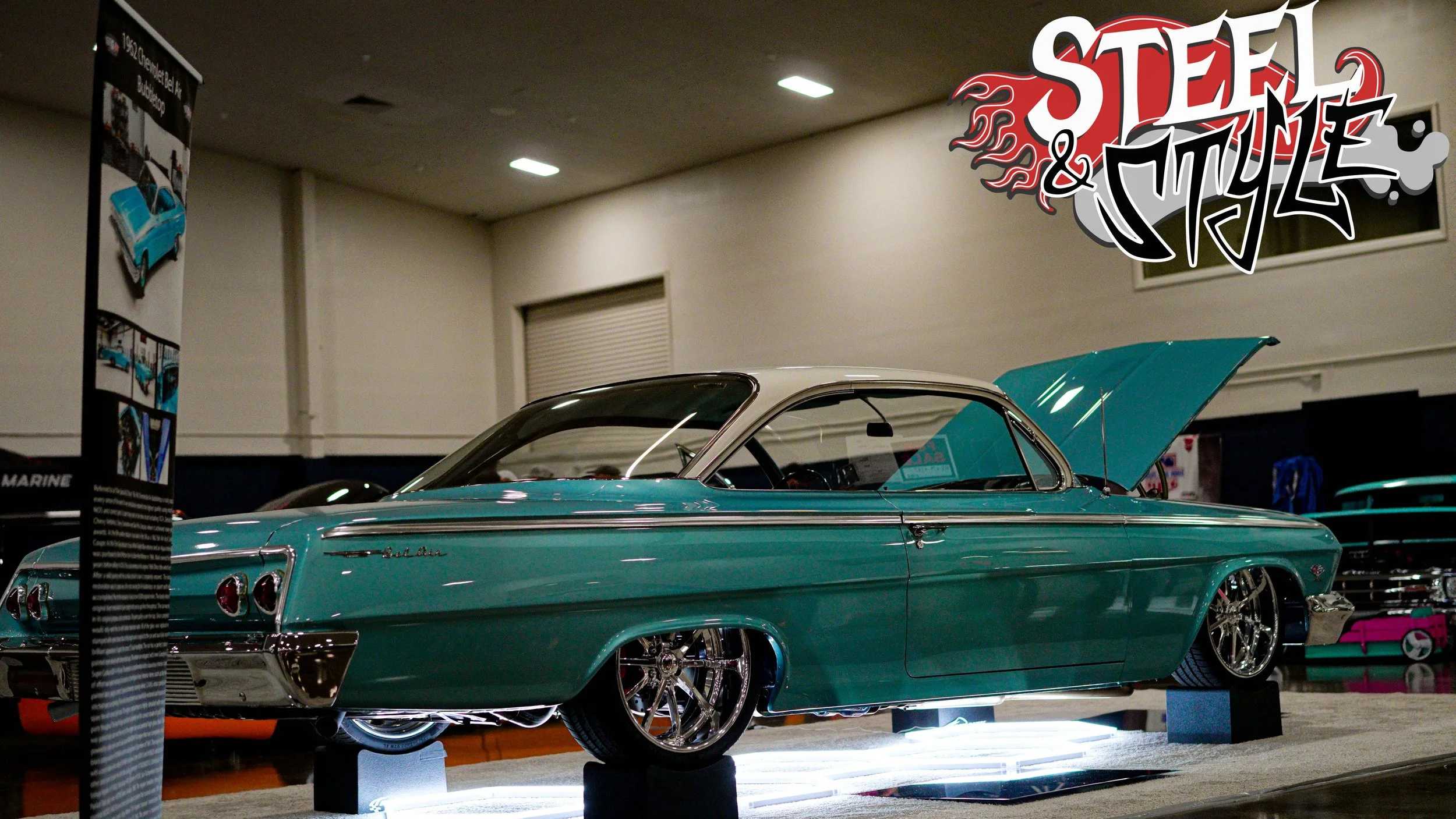 A vintage turquoise Chevrolet Bel Air car with the hood open on display at an indoor car show, with a 'Steel & Style' logo in the top right corner.