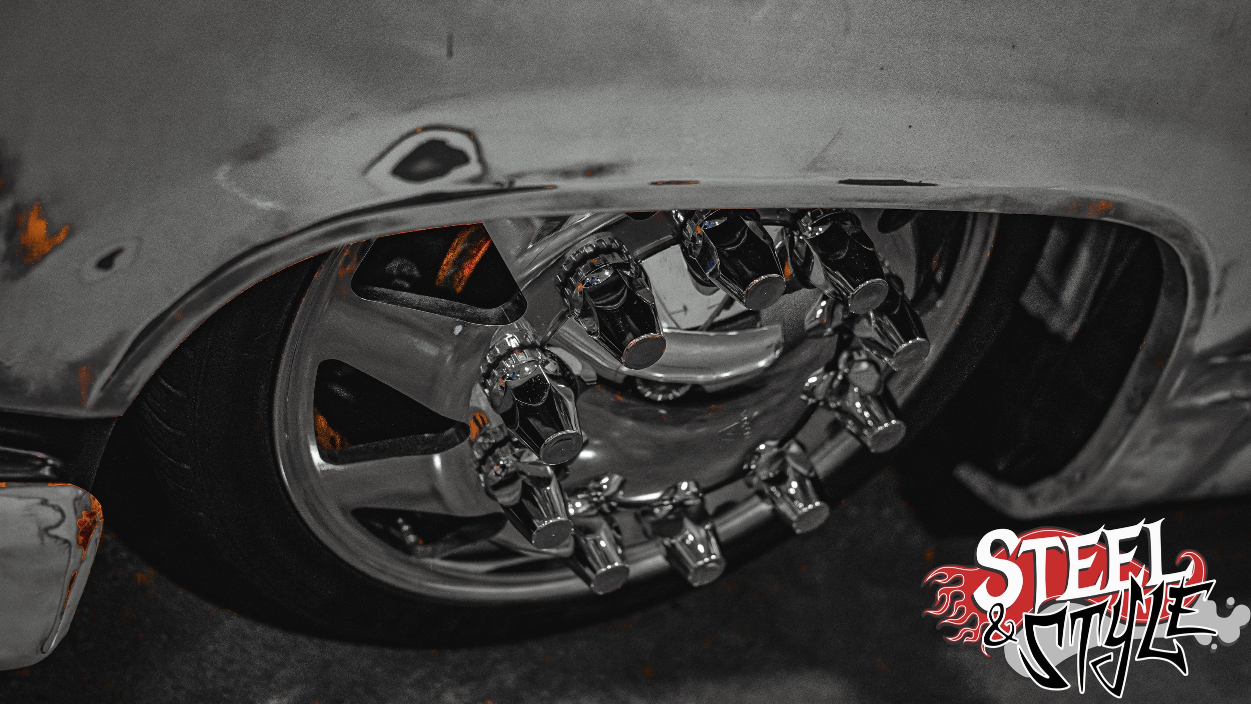 Close-up of a chrome wheel with multiple valve caps and a tire, part of a vintage or custom car, with a 'Steel & Style' logo in the bottom right corner.