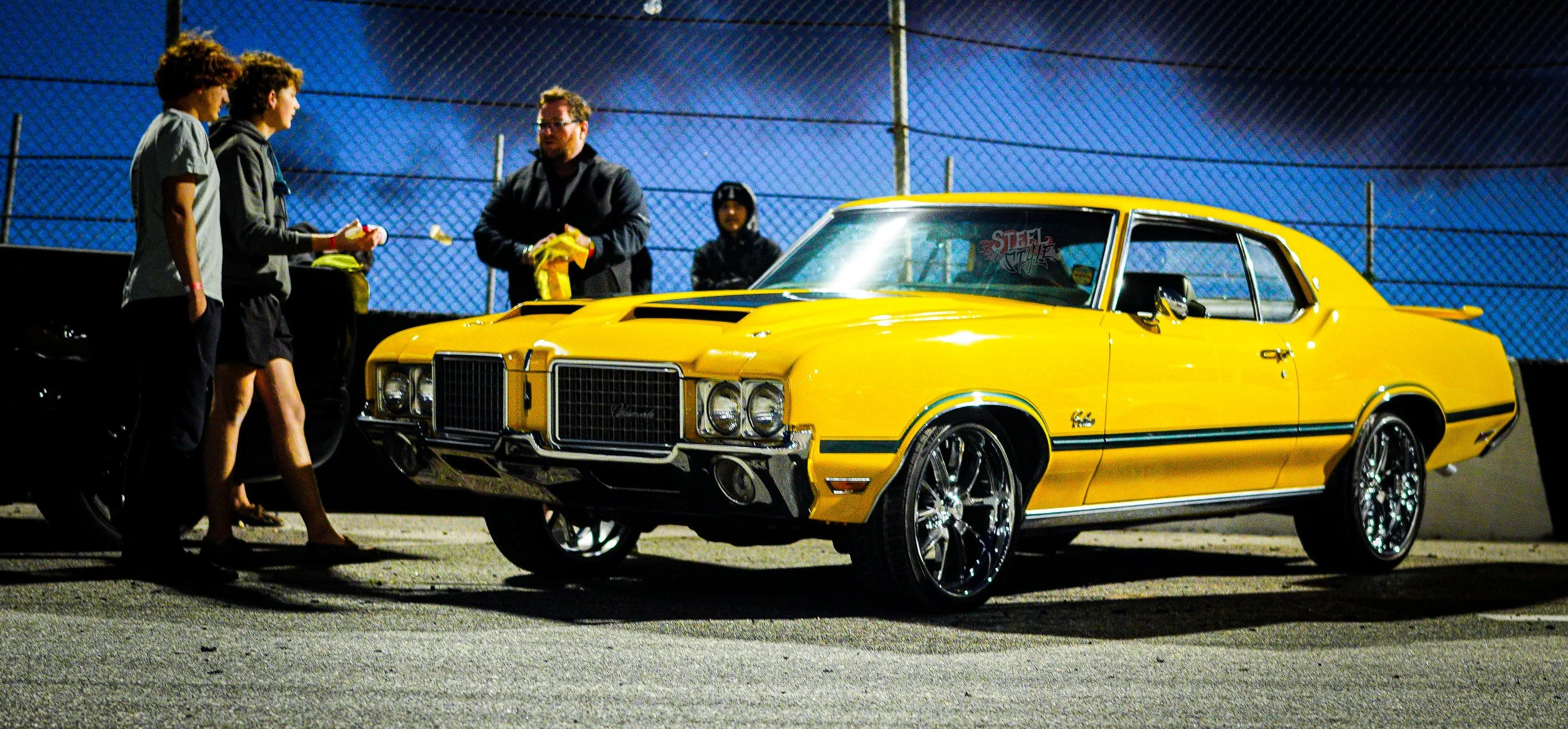 A yellow vintage muscle car with chrome rims parked on a dark street at night, people standing nearby, against a background of a chain-link fence and blue windscreens.