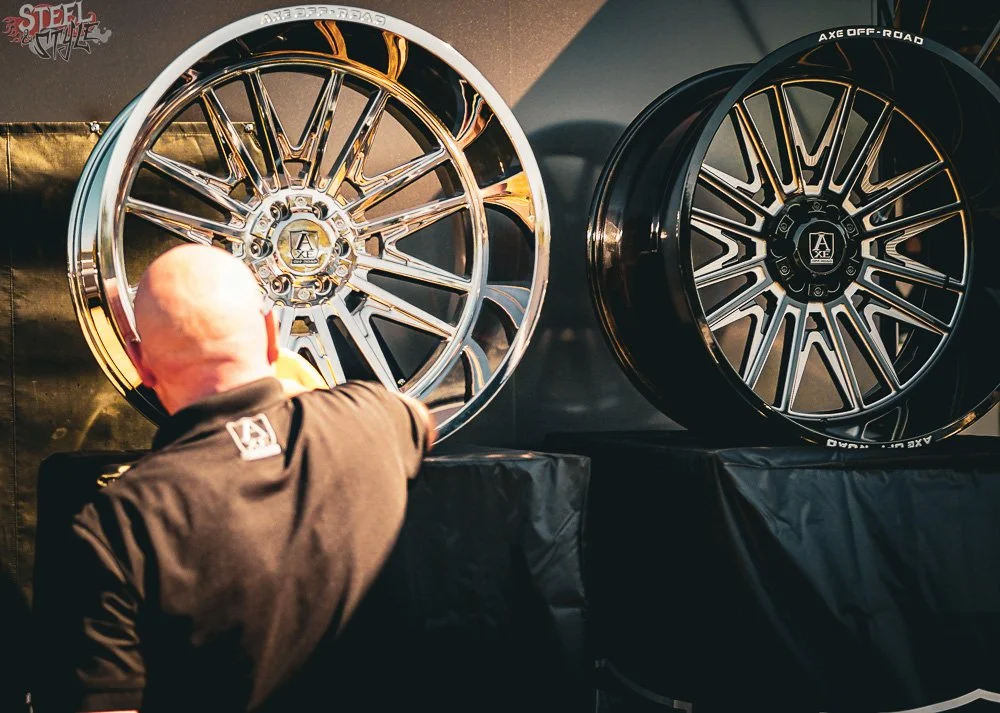 A person with a bald head and black shirt looking at two large, aftermarket alloy wheels on display. The wheels have intricate spoke designs, one polished chrome and the other black with machined accents, on a black display table.