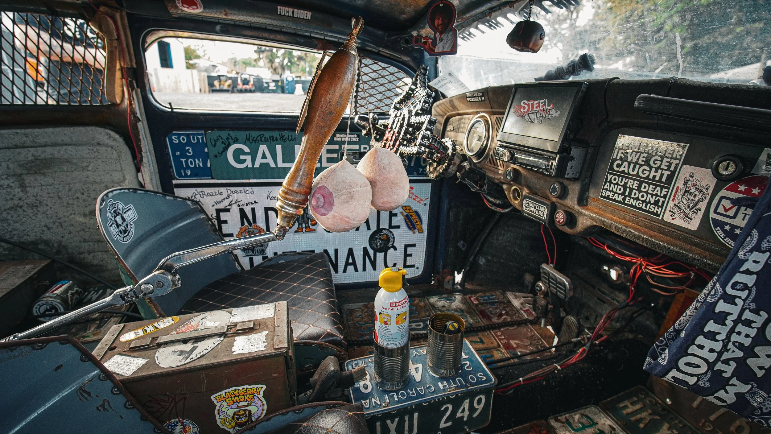 Interior of a decorated vehicle with various stickers, signs, and miscellaneous items, including a steering wheel, dashboard, and gear shift. Items like spray bottles, cans, and personal memorabilia are visible.