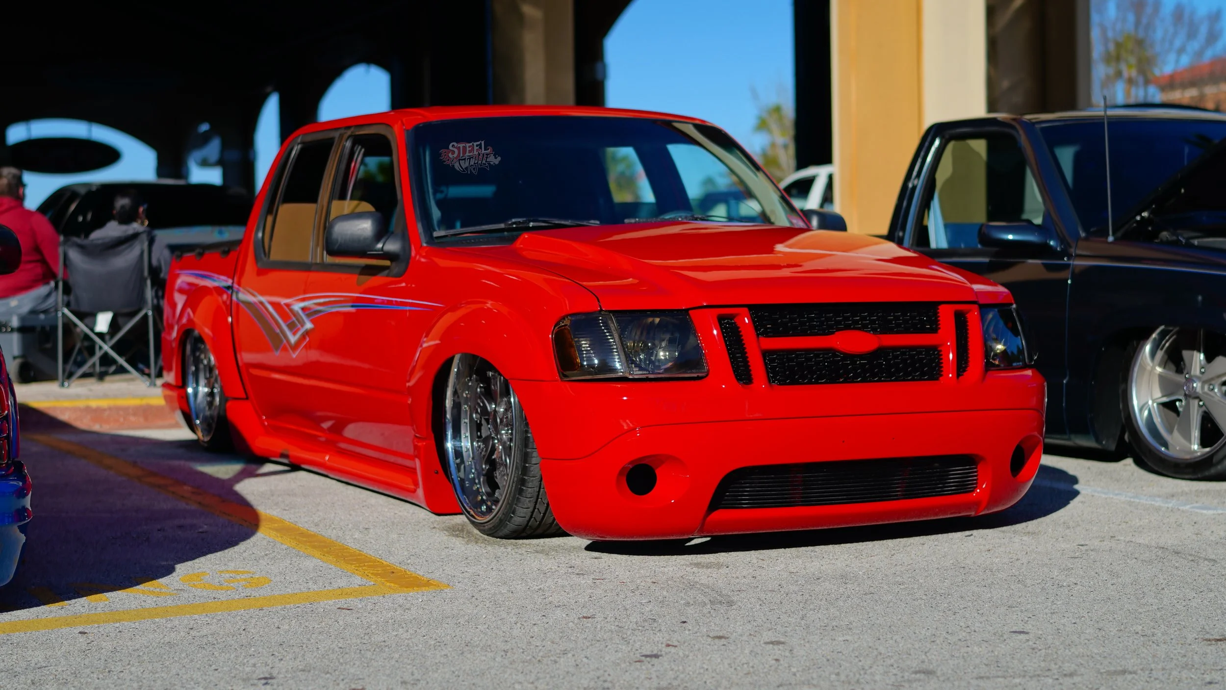 Red modified pickup truck with lowered suspension and custom wheels parked in an outdoor lot at a car show.