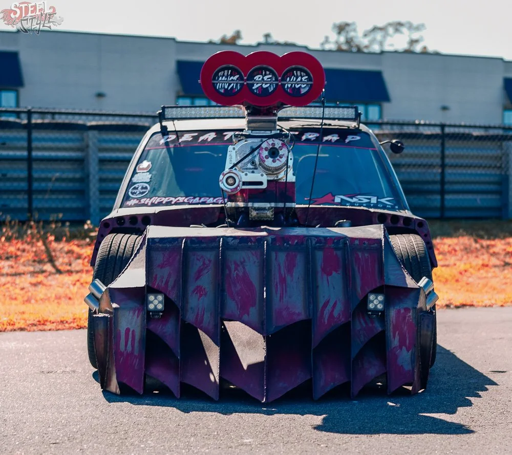 A custom race car with a large front splitter, oversized rear wing, and racing decals, parked outdoors on asphalt with a building and chain-link fence in the background.