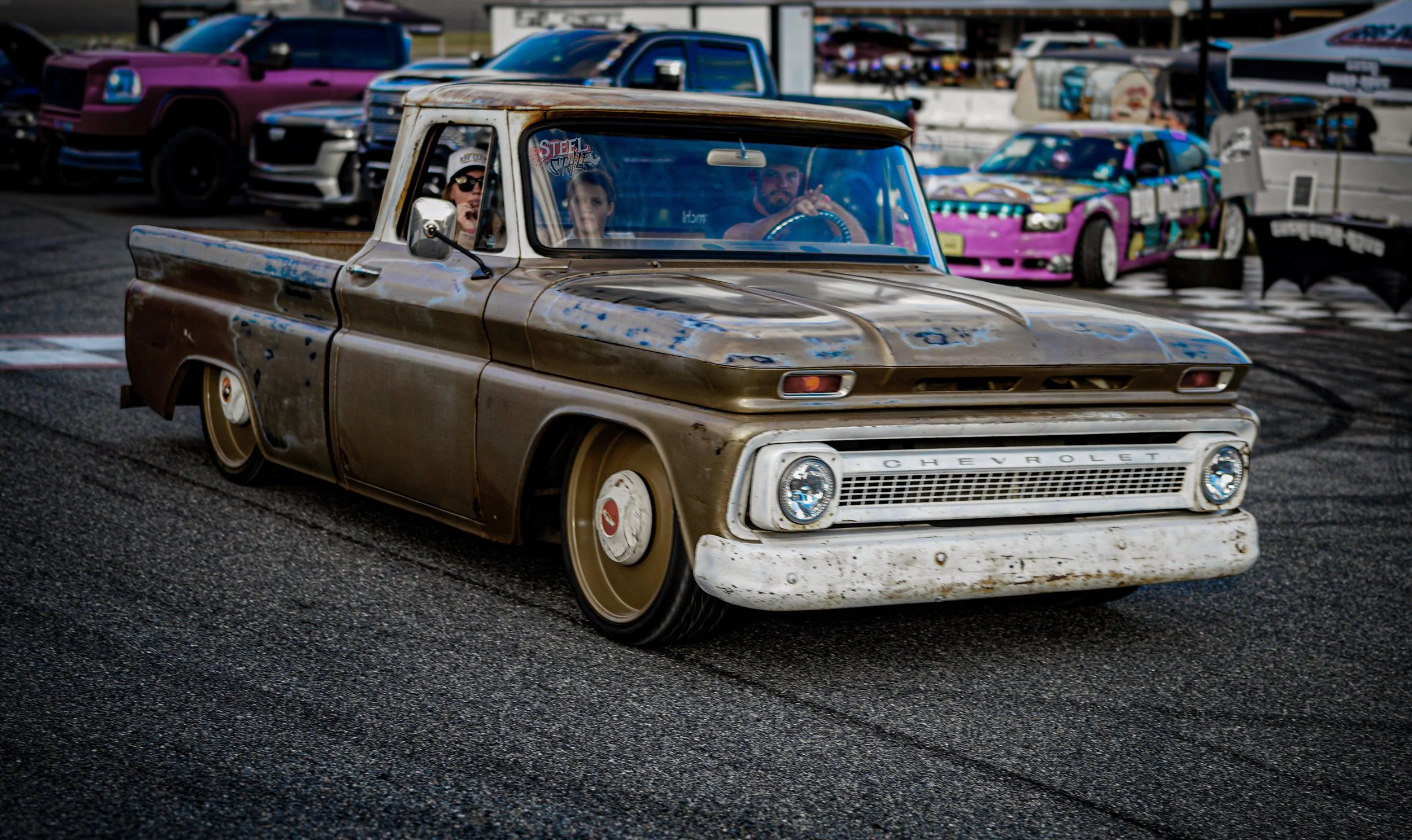 A custom vintage Chevrolet pickup truck with a rusty, weathered appearance drifting on a race track, with car enthusiasts and colorful race cars in the background.