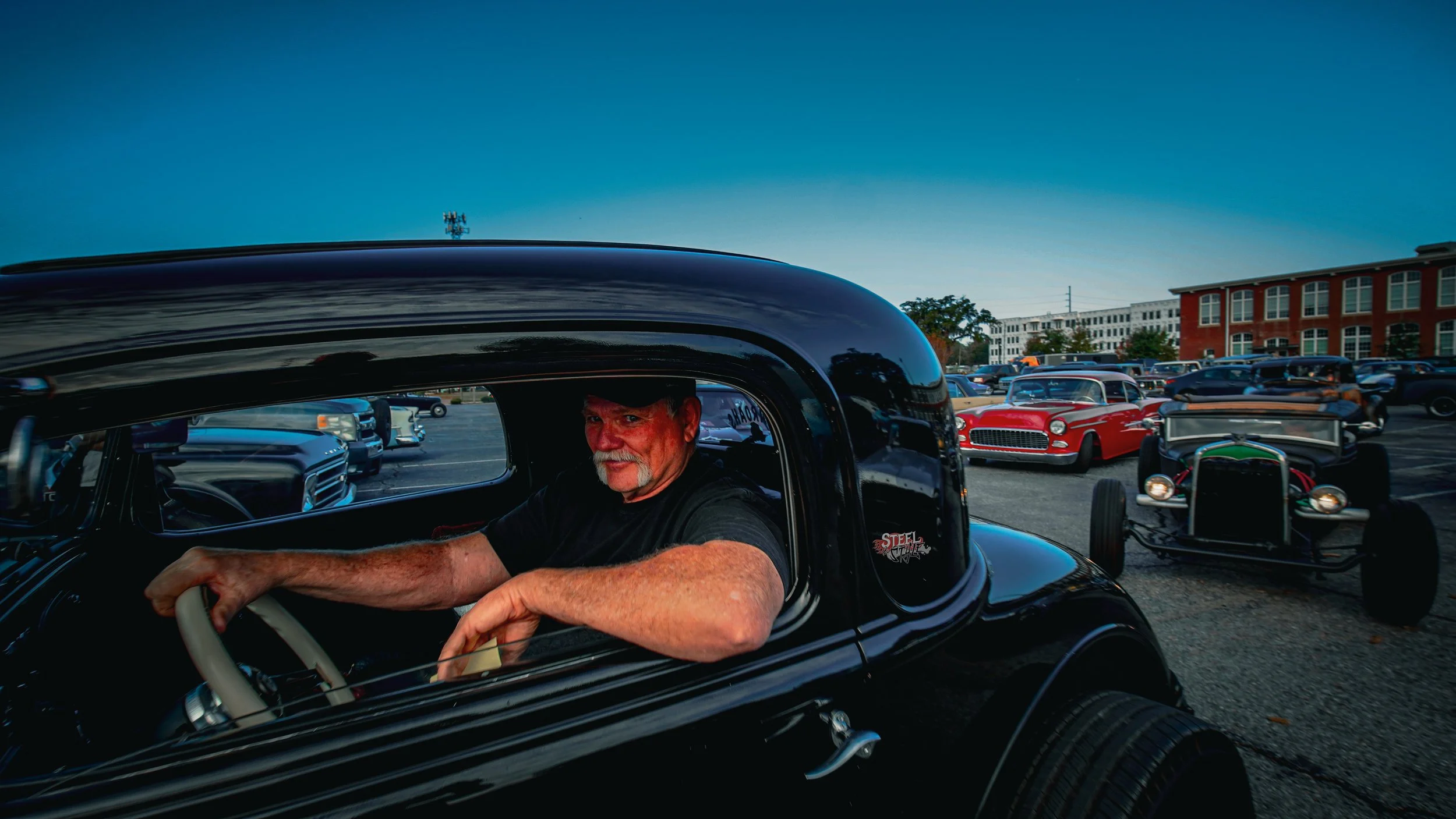 A man with a white beard and mustache sitting in the driver's seat of a black vintage car at a classic car show, with other vintage cars and a building in the background during evening.