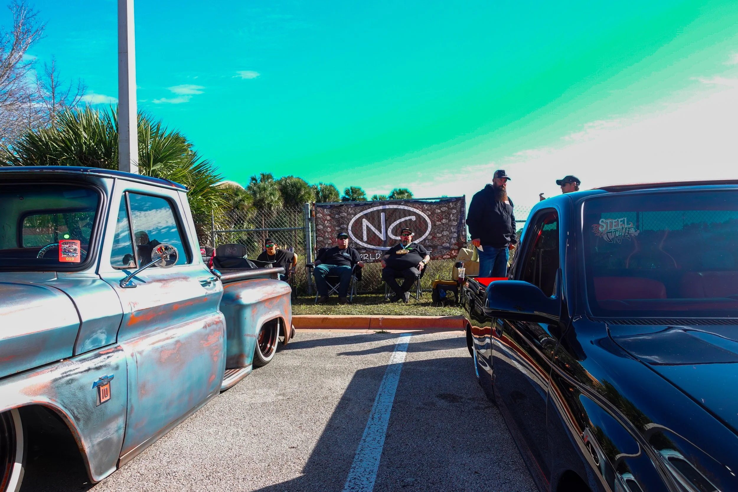 A car meet in a parking lot with two classic trucks, six men sitting or standing near a banner, and a clear sky with trees in the background.