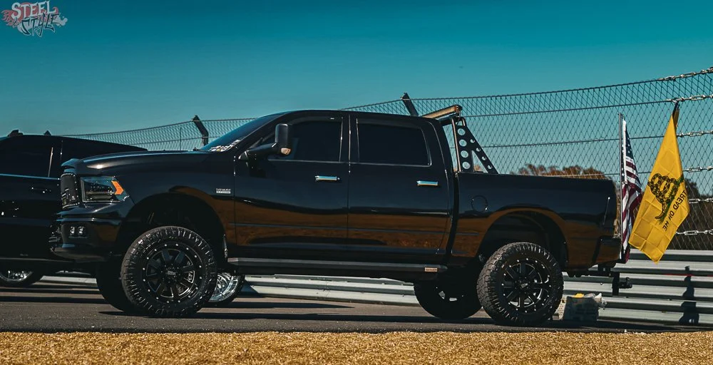 Black pickup truck with large tires and black rims parked on a race track, flags flapping behind it, under a cloudy sky.