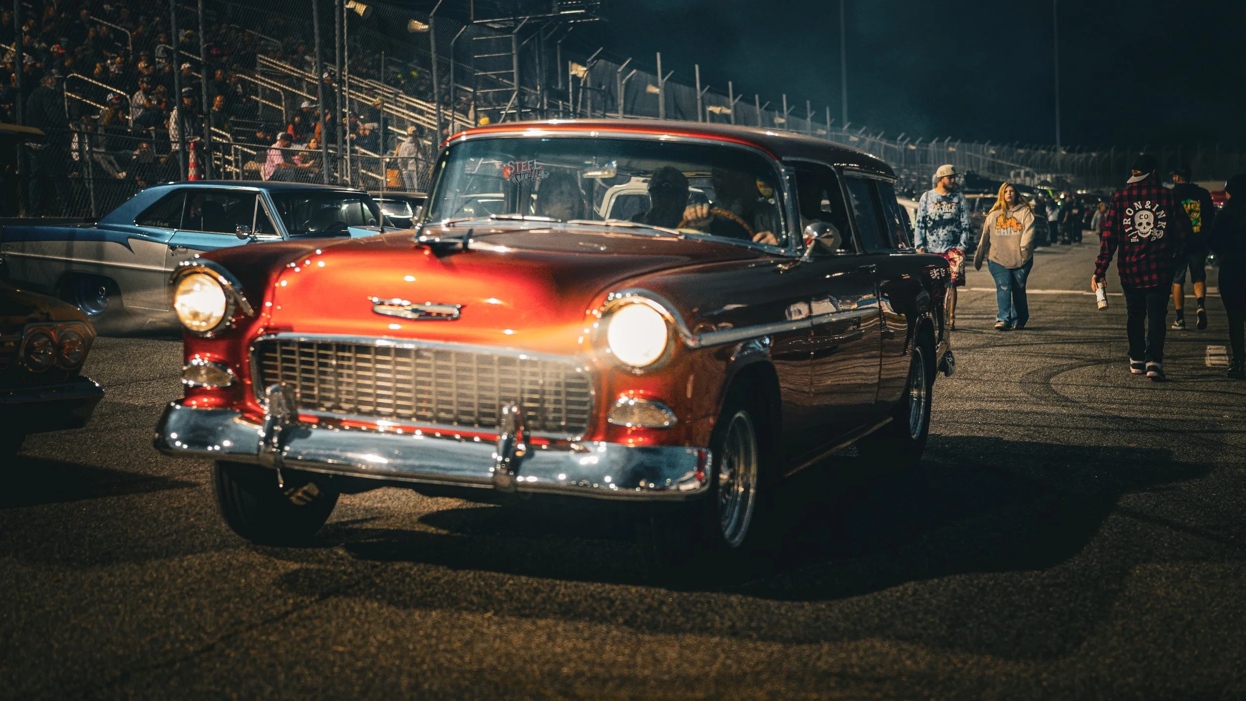 A vintage red and black car parked at night with a crowd of people and other classic cars in the background.
