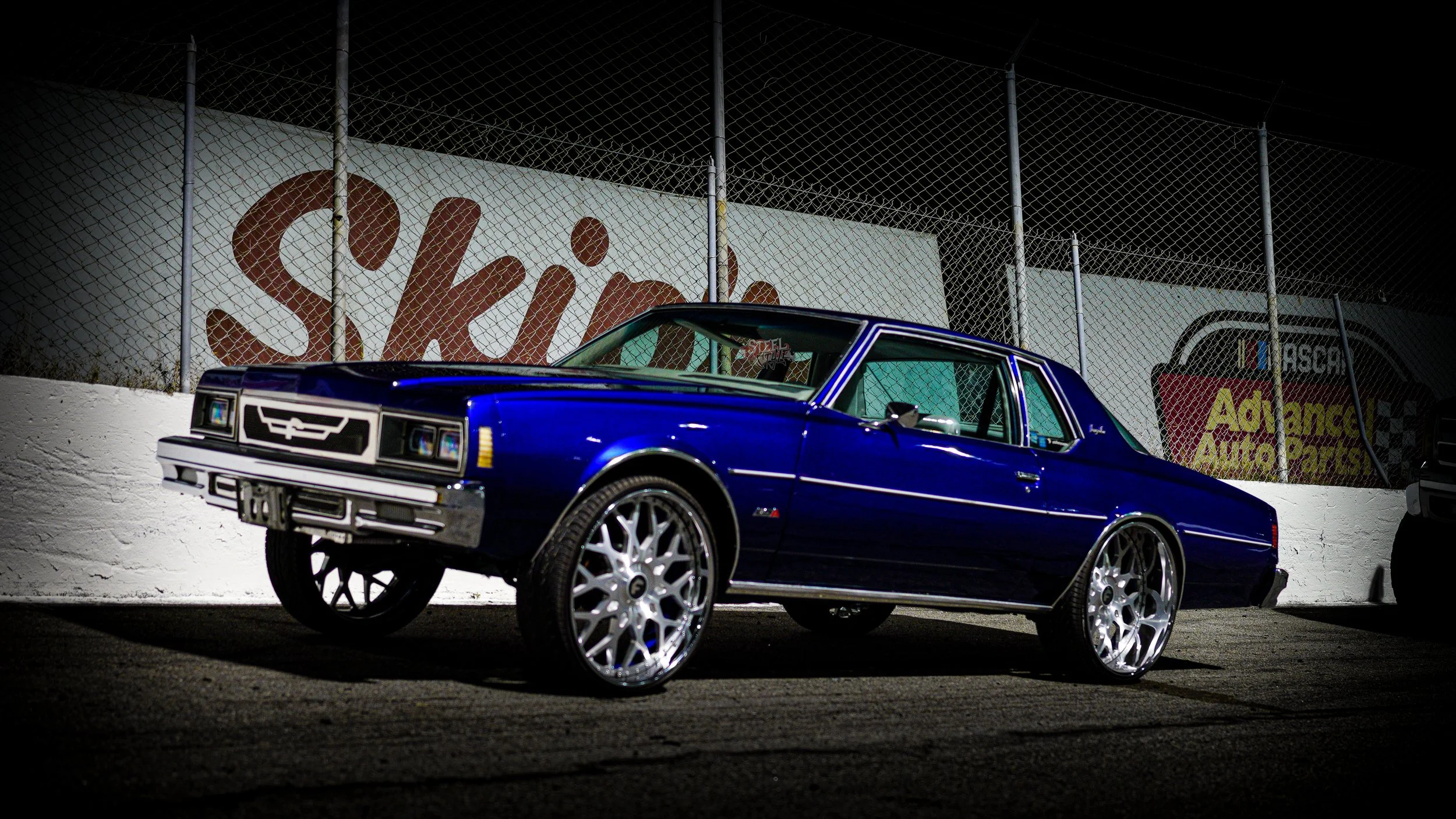 A vintage blue Chevrolet car with large silver wheels parked on a racetrack at night, chain-link fence and signs in the background.