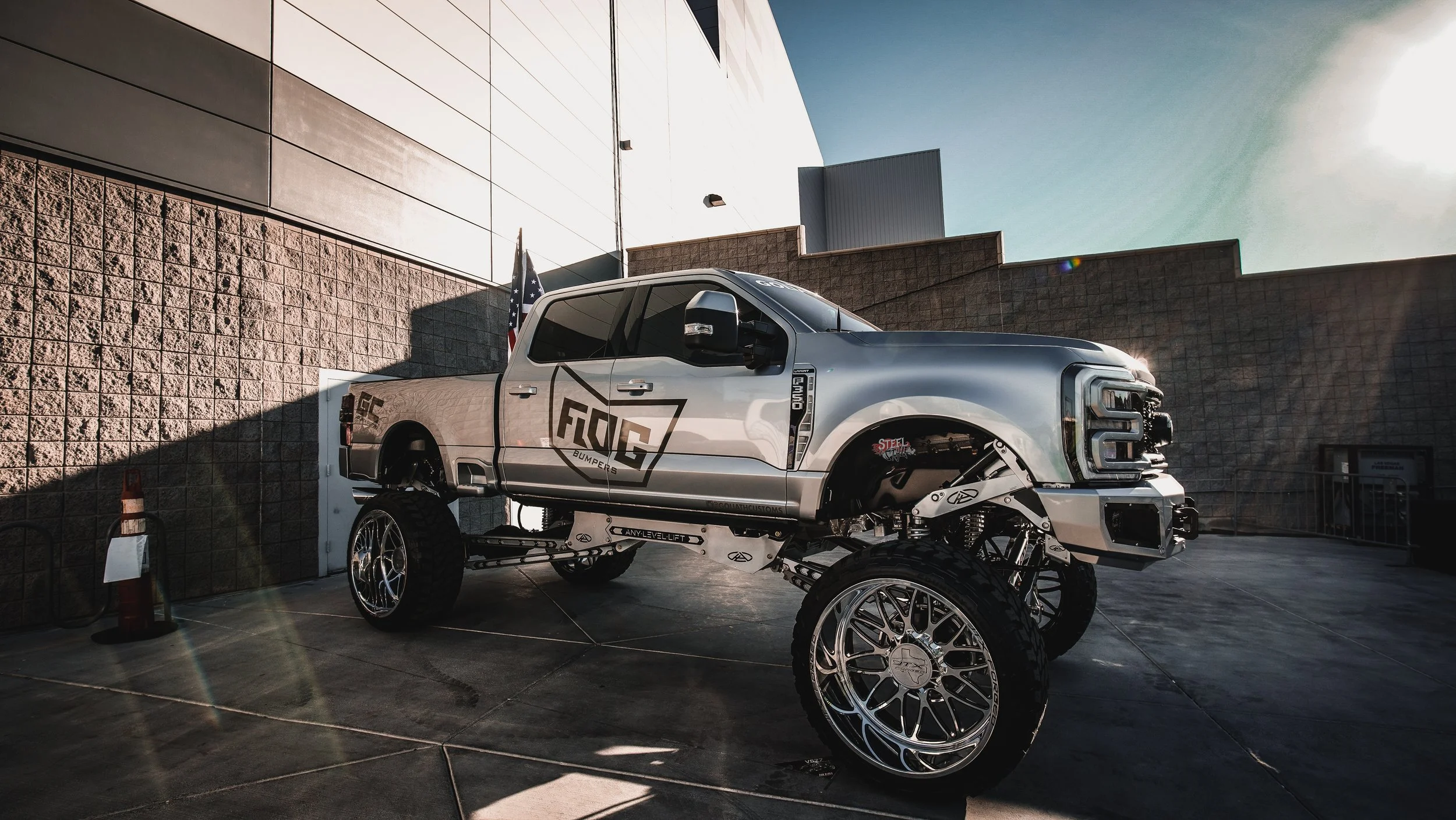 A custom lifted pickup truck with oversized chrome wheels, logo on the side, and no front wheels, parked outdoors near a brick and stucco wall during sunset.