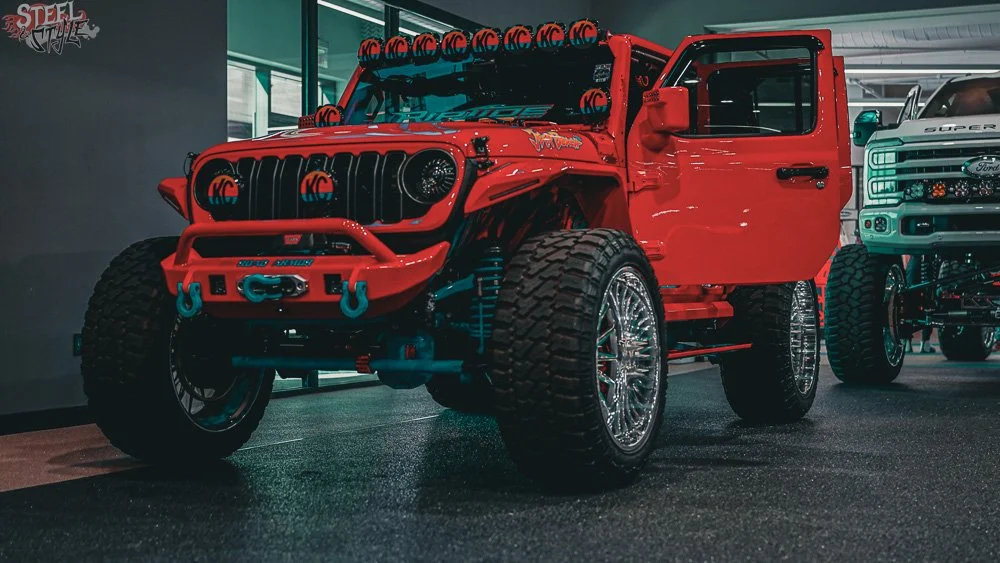 A modified red off-road Jeep with large tires, custom bumpers, multiple KC LED lights on the roof, and chrome wheels, displayed indoors at a car show.