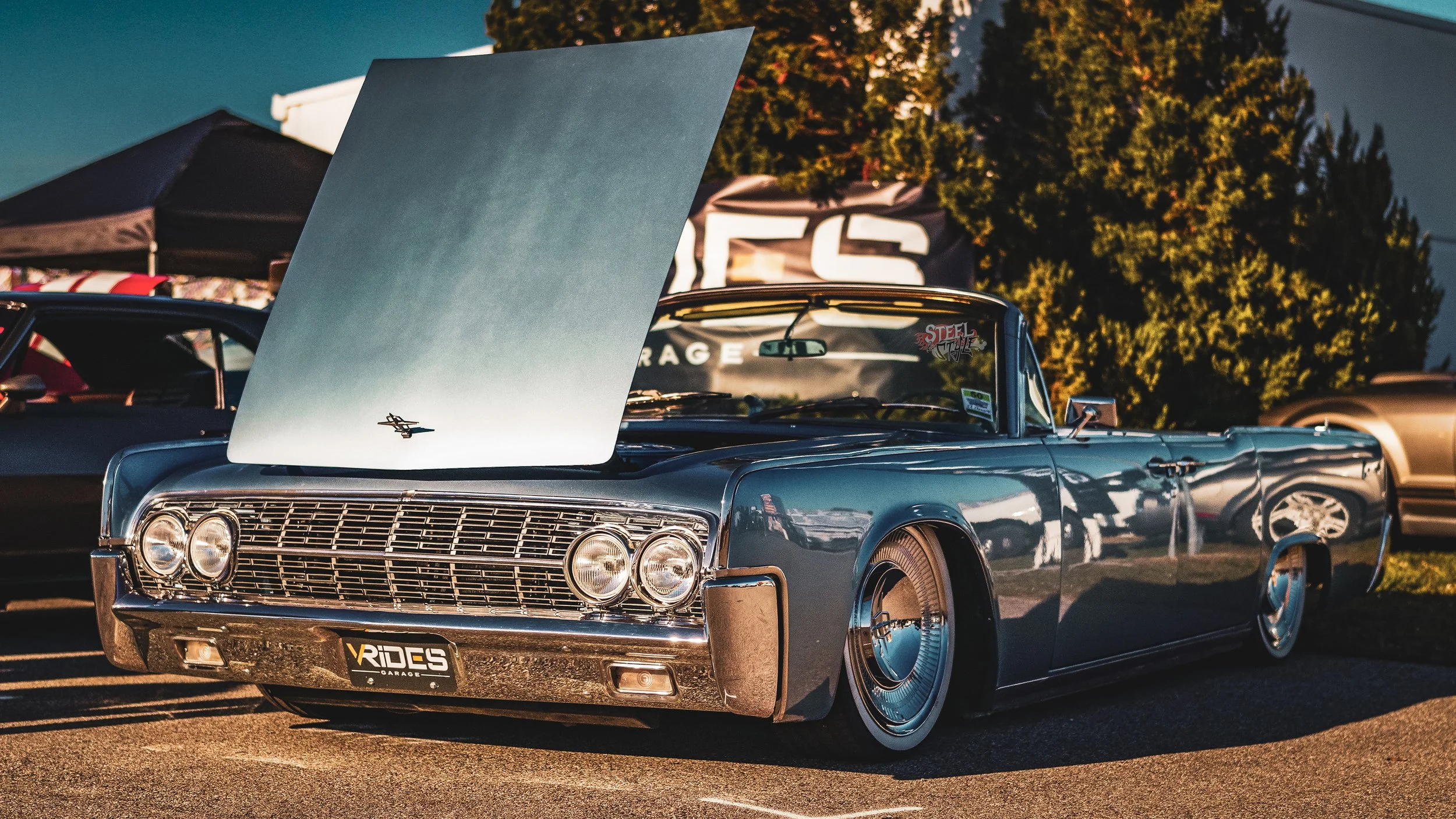 A vintage blue lowrider car with its hood open at an outdoor car show during sunset, surrounded by other cars and trees in the background.