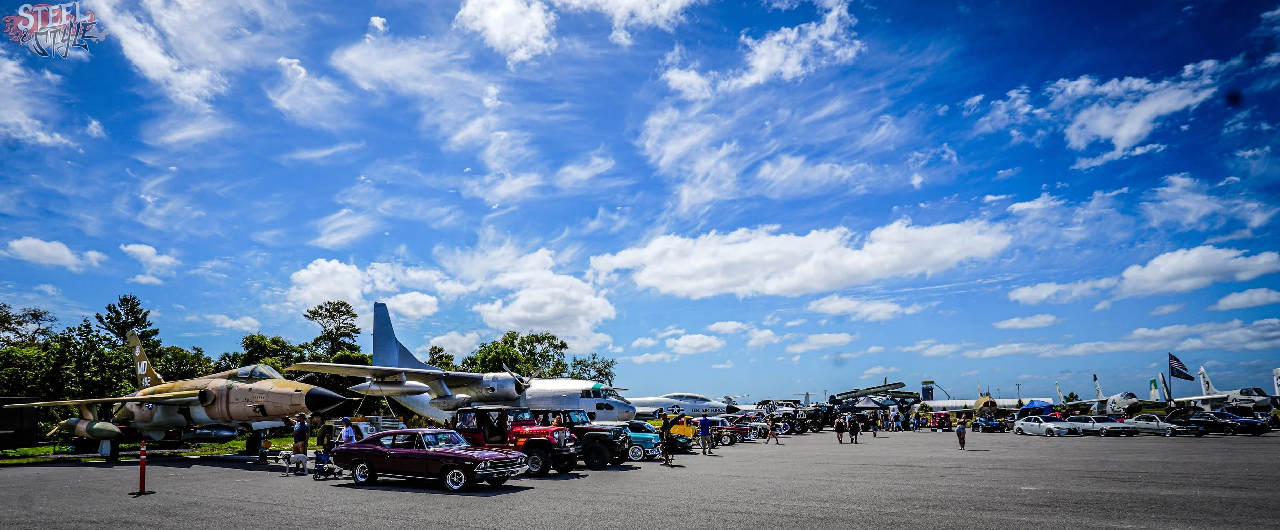 Outdoor aircraft and vintage cars displayed at an air show with a bright blue sky and scattered clouds.