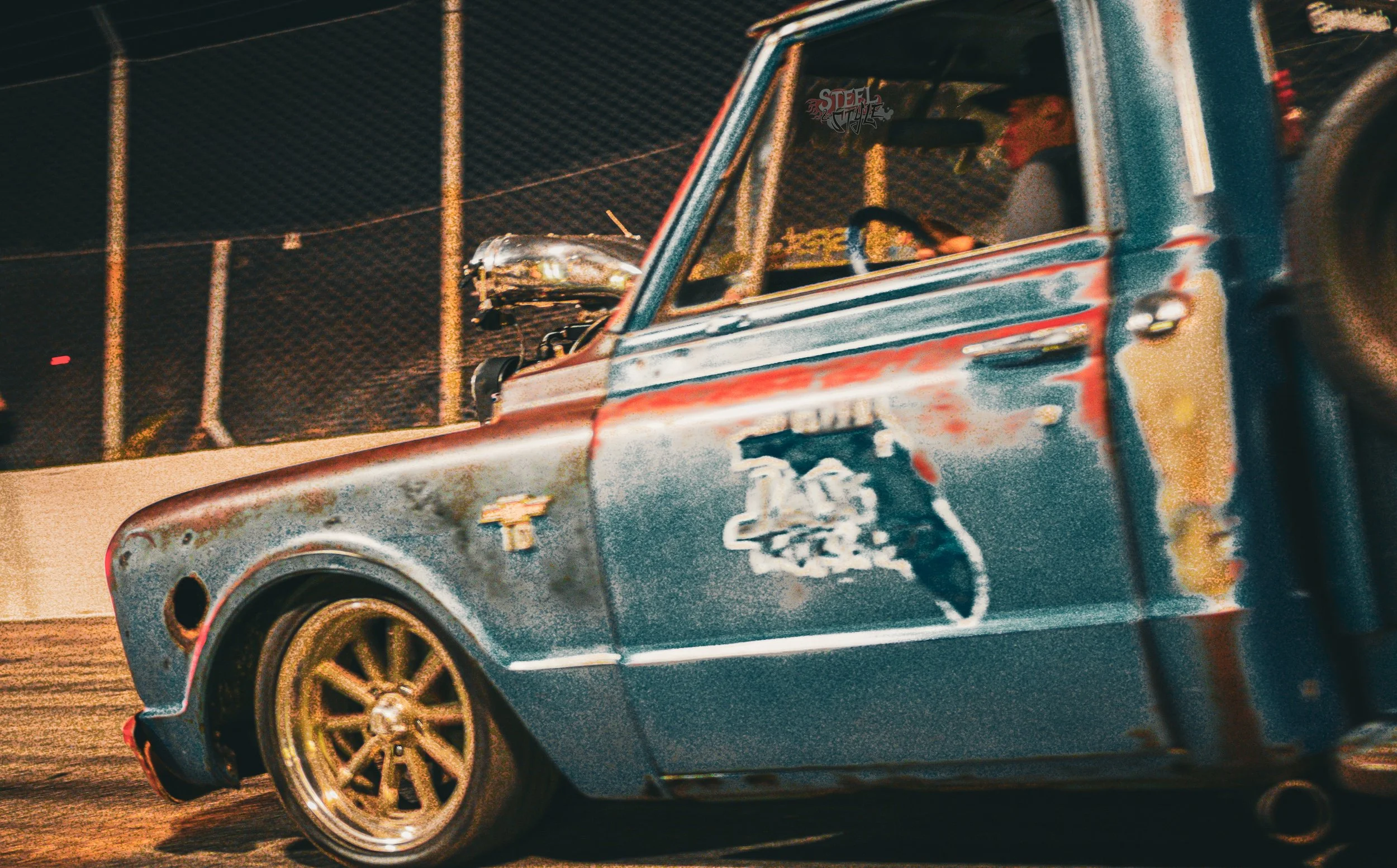Close-up of a vintage race car with a weathered, rusted blue exterior, gold wheels, and a racing number, parked on a dirt track at night, with a driver inside and chain-link fence in the background.