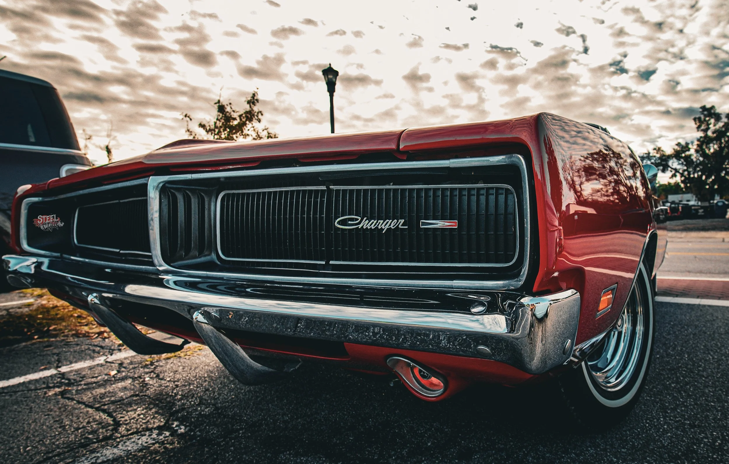 Front view of a red vintage Dodge Charger car parked on an asphalt lot under a cloudy sky, with trees and other vehicles in the background.