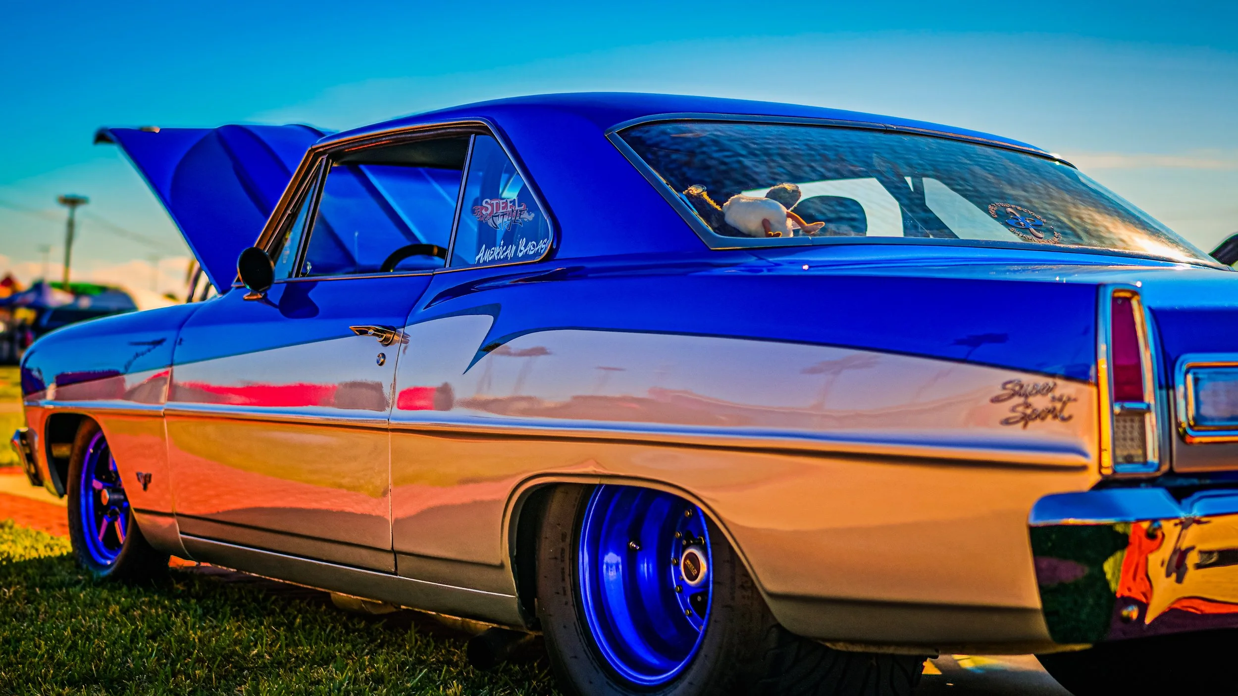 A vintage blue and silver super sport car on a grassy field at sunset, with open hood and custom wheels.