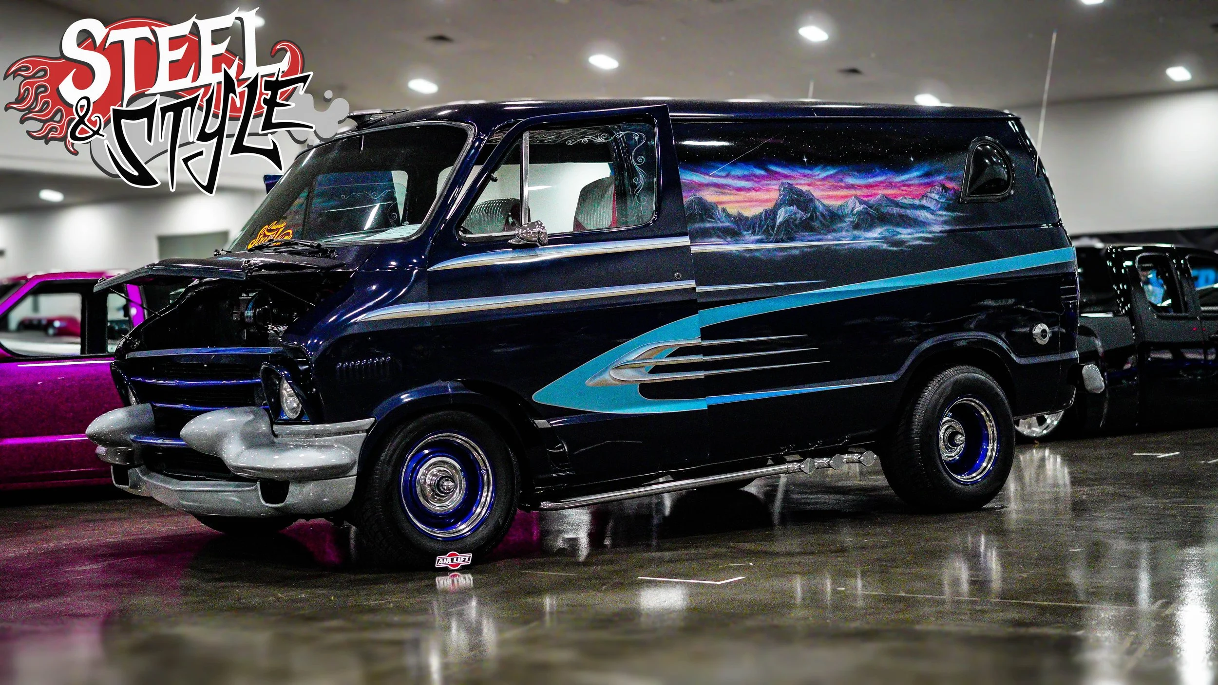 A vintage van with custom paint, featuring a mountain landscape and blue accents, parked indoors at a car show, with other vehicles in the background.