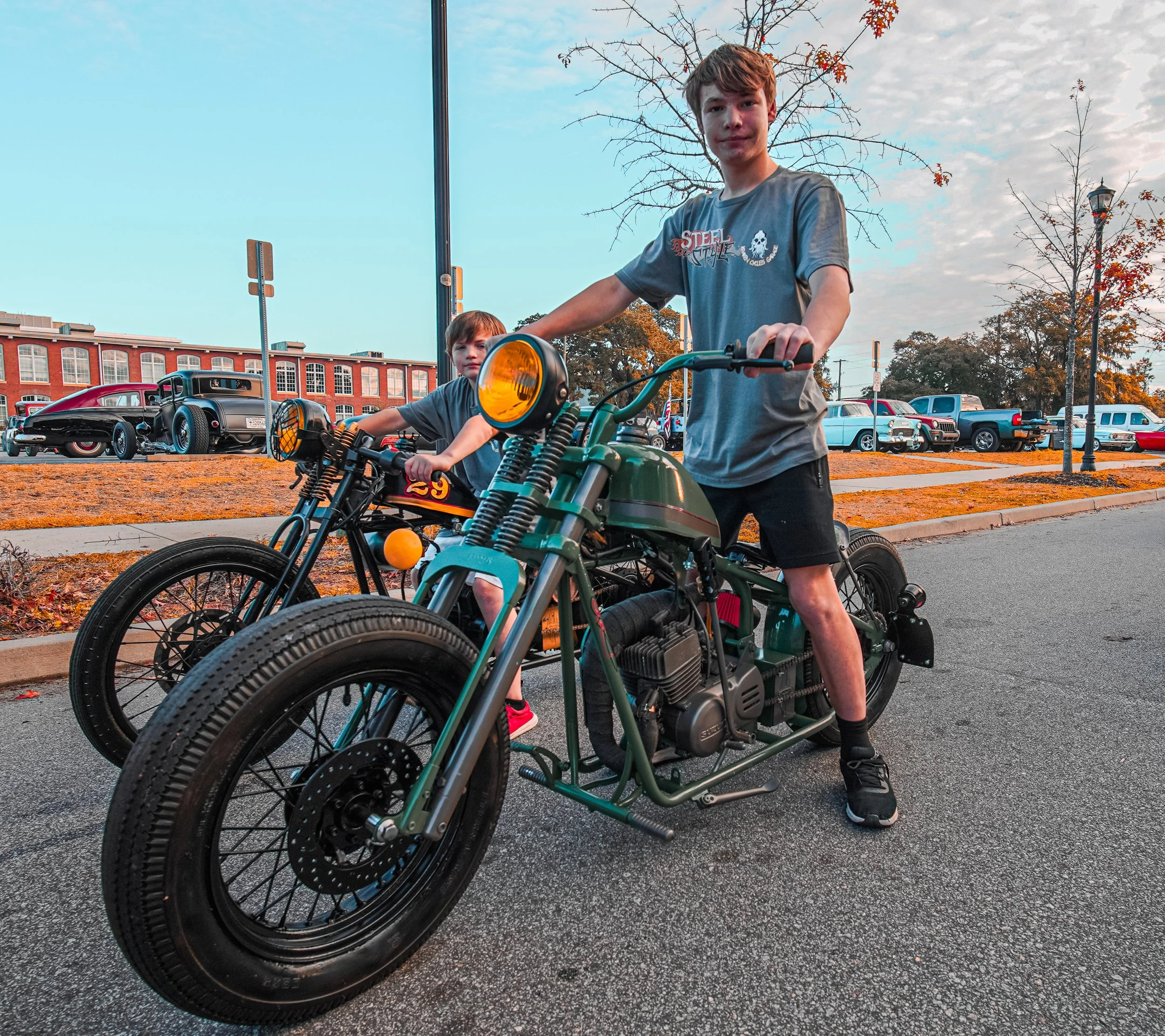Two boys with bicycles on a street during the daytime, one on a vintage motorcycle and the other on a smaller bicycle, with a red brick building and parked cars in the background.
