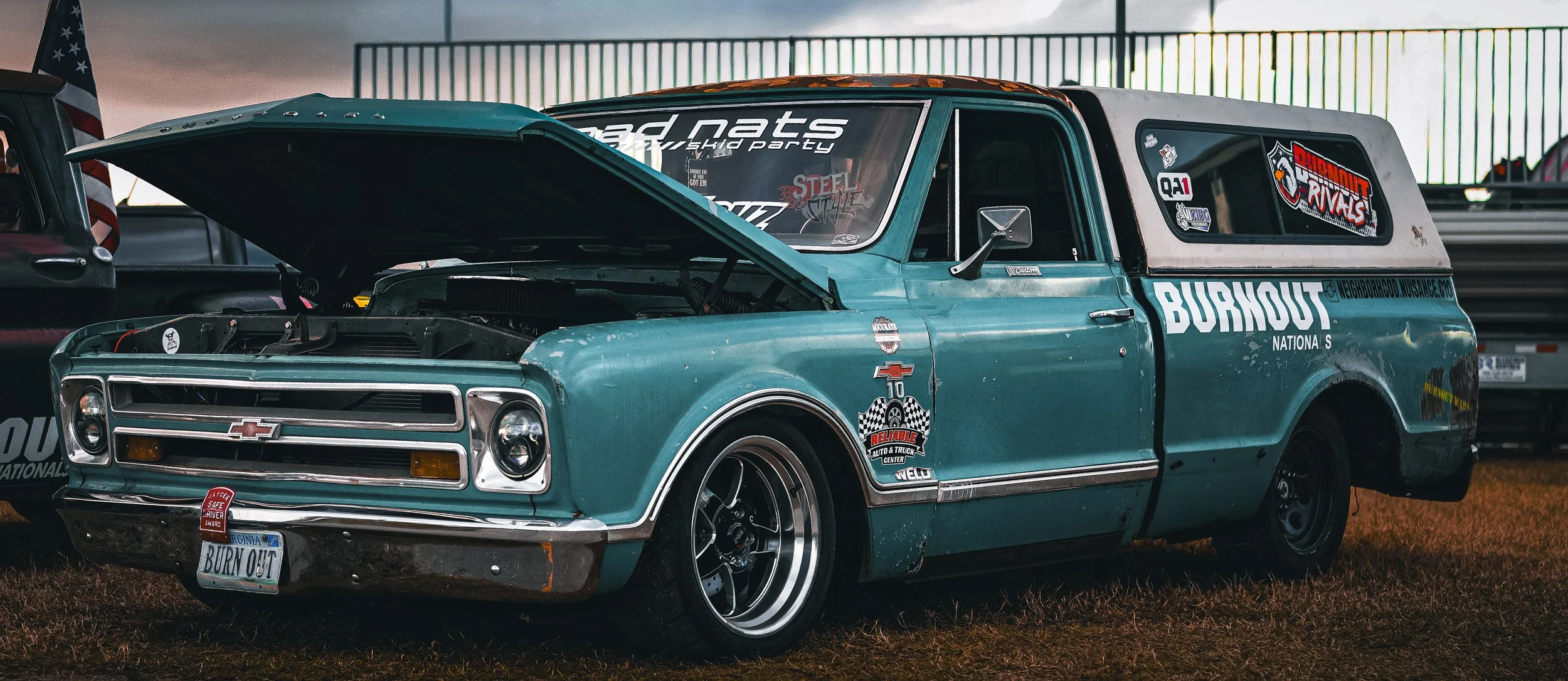 A vintage teal Chevrolet pickup truck with an open hood, racing decals, and custom wheels, parked on grass during the day.
