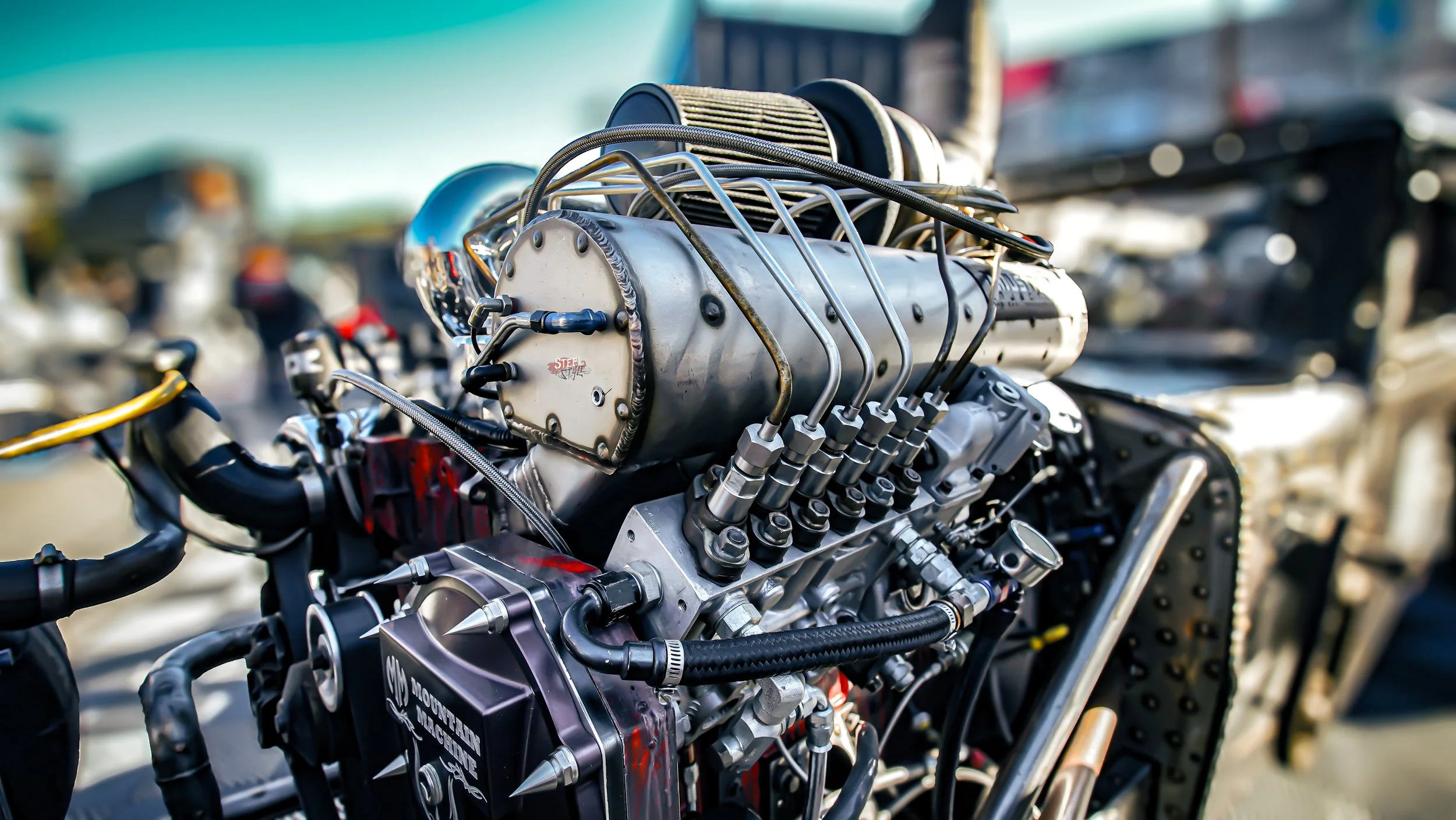 Close-up of a high-performance racing engine with various metallic components, hoses, and fittings, mounted on a vehicle in a racing paddock.