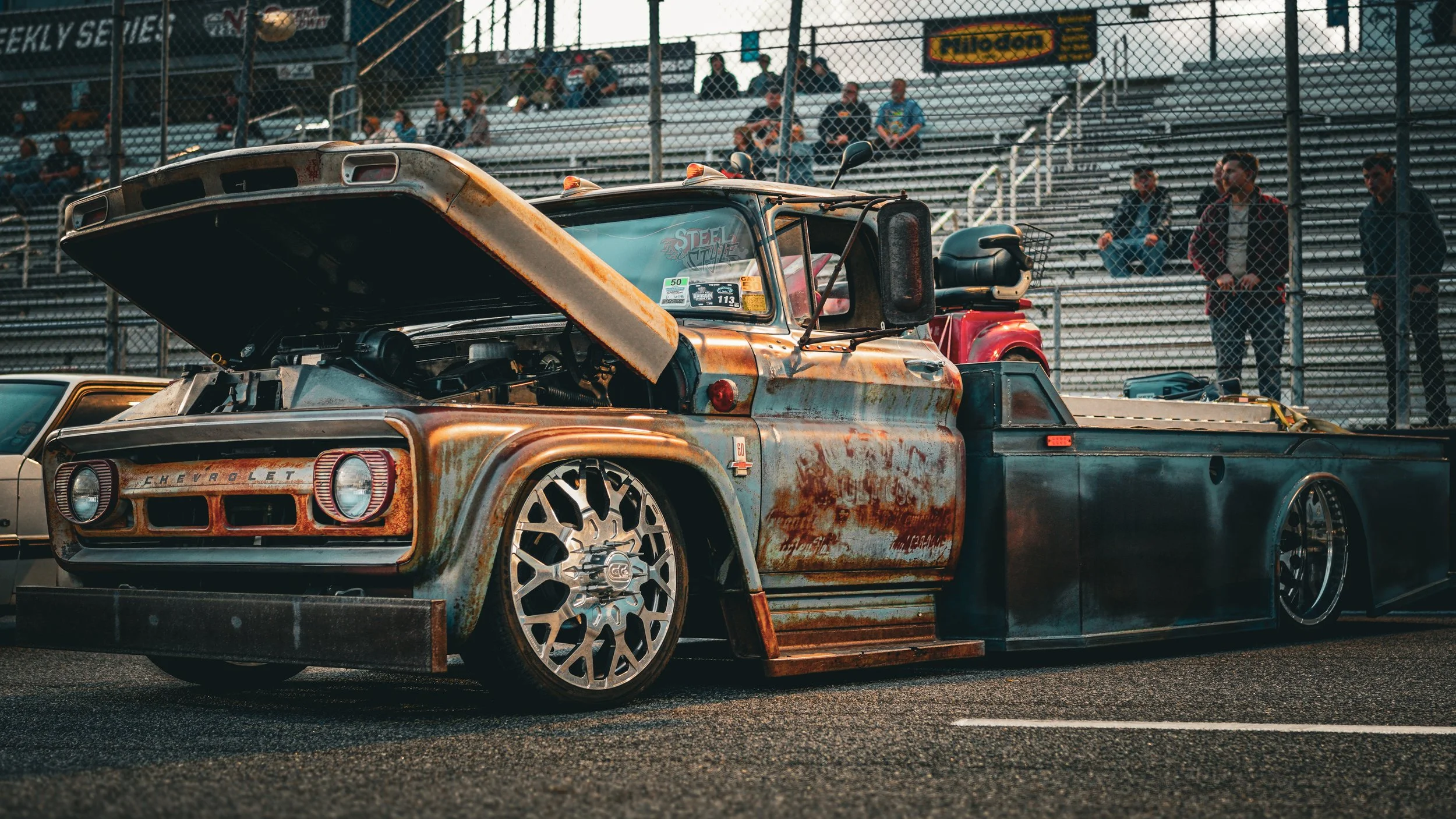 Rusty custom vintage Chevrolet pickup truck with lowered suspension and large alloy wheels at a car show. People sit in bleachers behind a fence in the background.