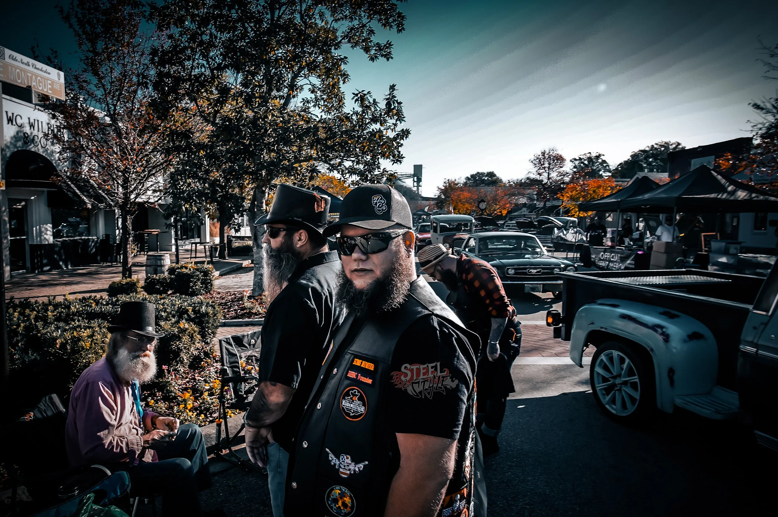 A group of men with beards dressed in biker attire, wearing sunglasses and hats, standing outdoors among vintage cars and market stalls in a town square during late afternoon or early evening, with trees displaying fall foliage.