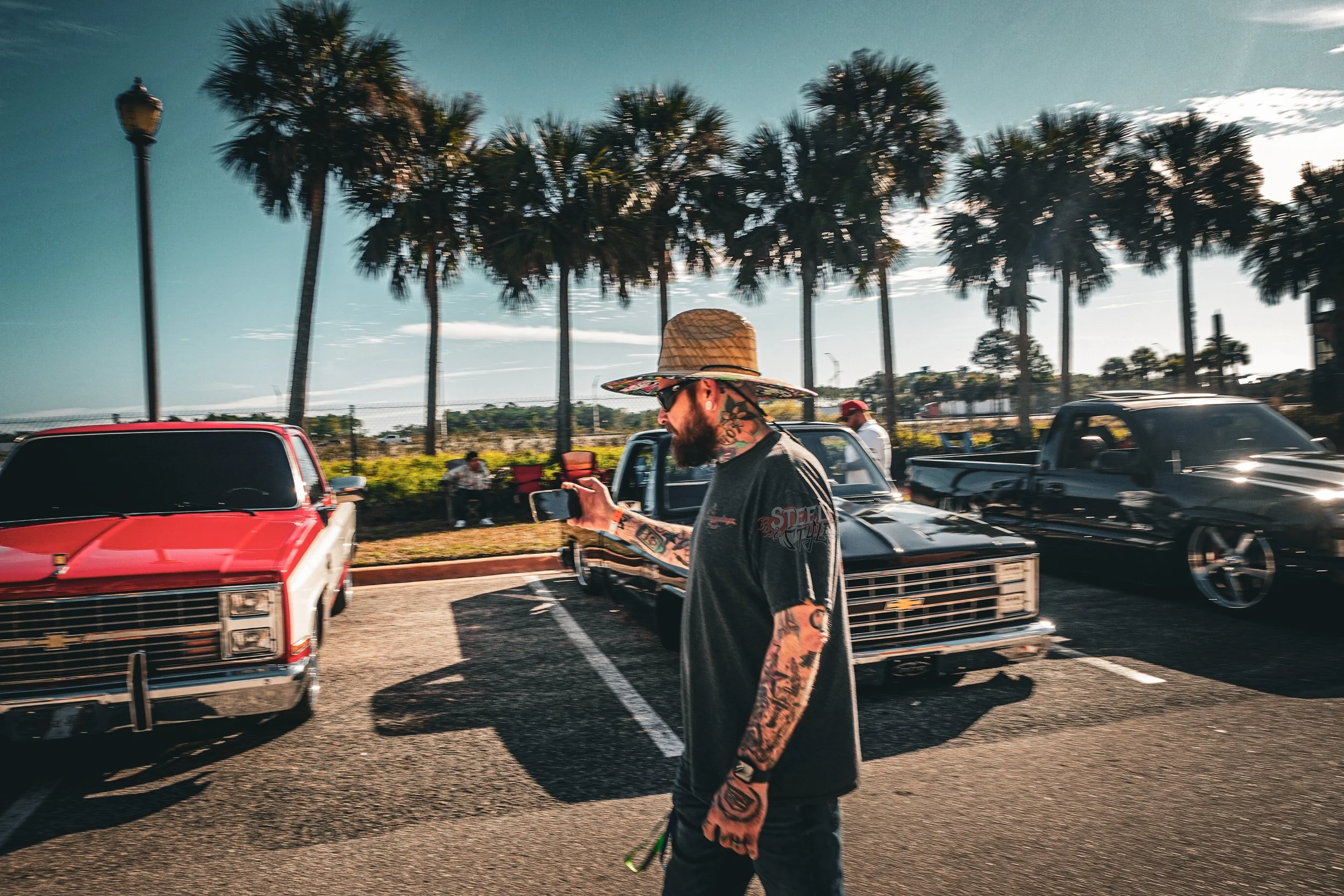 A man with tattoos and a beard wearing a wide-brimmed straw hat, sunglasses, a black graphic T-shirt, and jeans, walking in a parking lot while looking at his phone. Cars and palm trees are in the background.