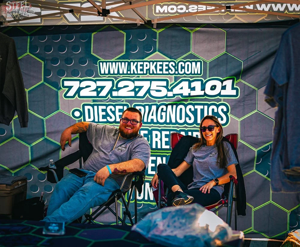 Two people sitting in camping chairs at an event booth, smiling. The booth has a large promotional banner in the background with a website and phone number for diesel diagnostics and repair.