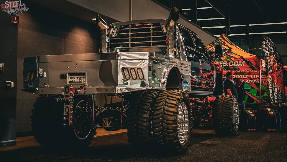 A customized monster truck with oversized tires and metallic body panels on display indoors, featuring a flag, promotional banners, and people nearby.