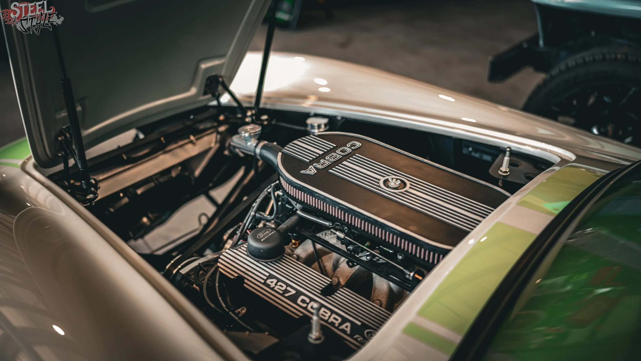 Close-up of a classic car's engine bay, showing a Cobra V8 engine with black and silver striped valve covers, and the hood open.