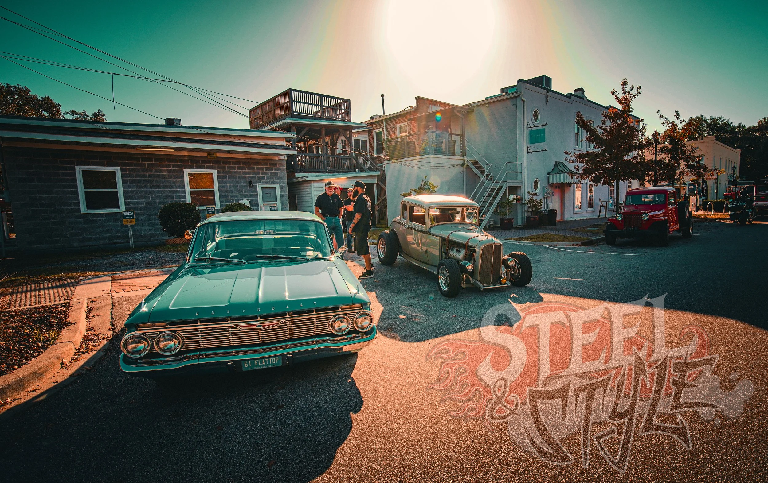Vintage cars parked on a street during sunset with a small group of people standing near the cars and colorful buildings in the background.