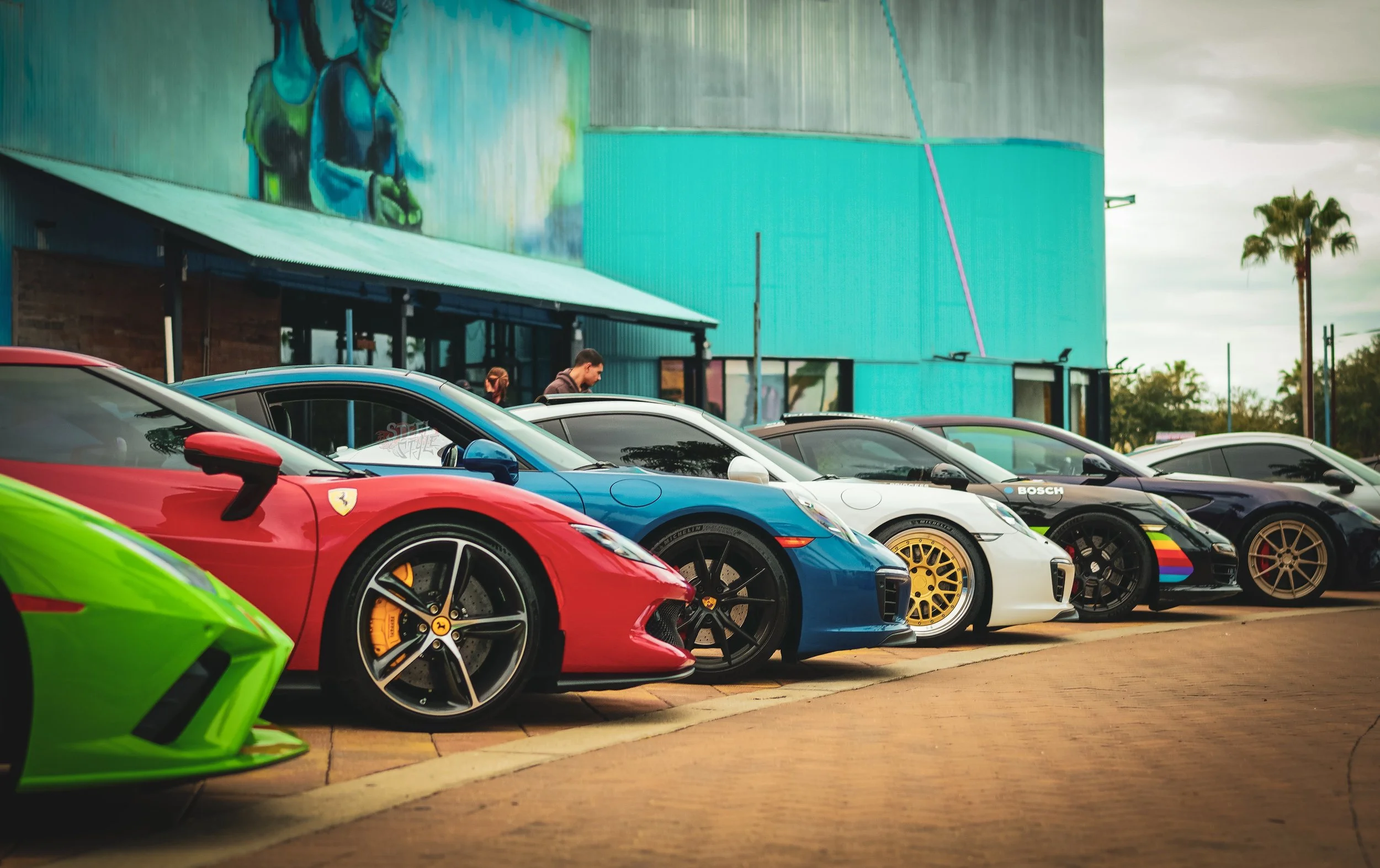 Lineup of colorful luxury sports cars parked in front of a building with graffiti art, including a green, red, blue, white, black, and purple car.