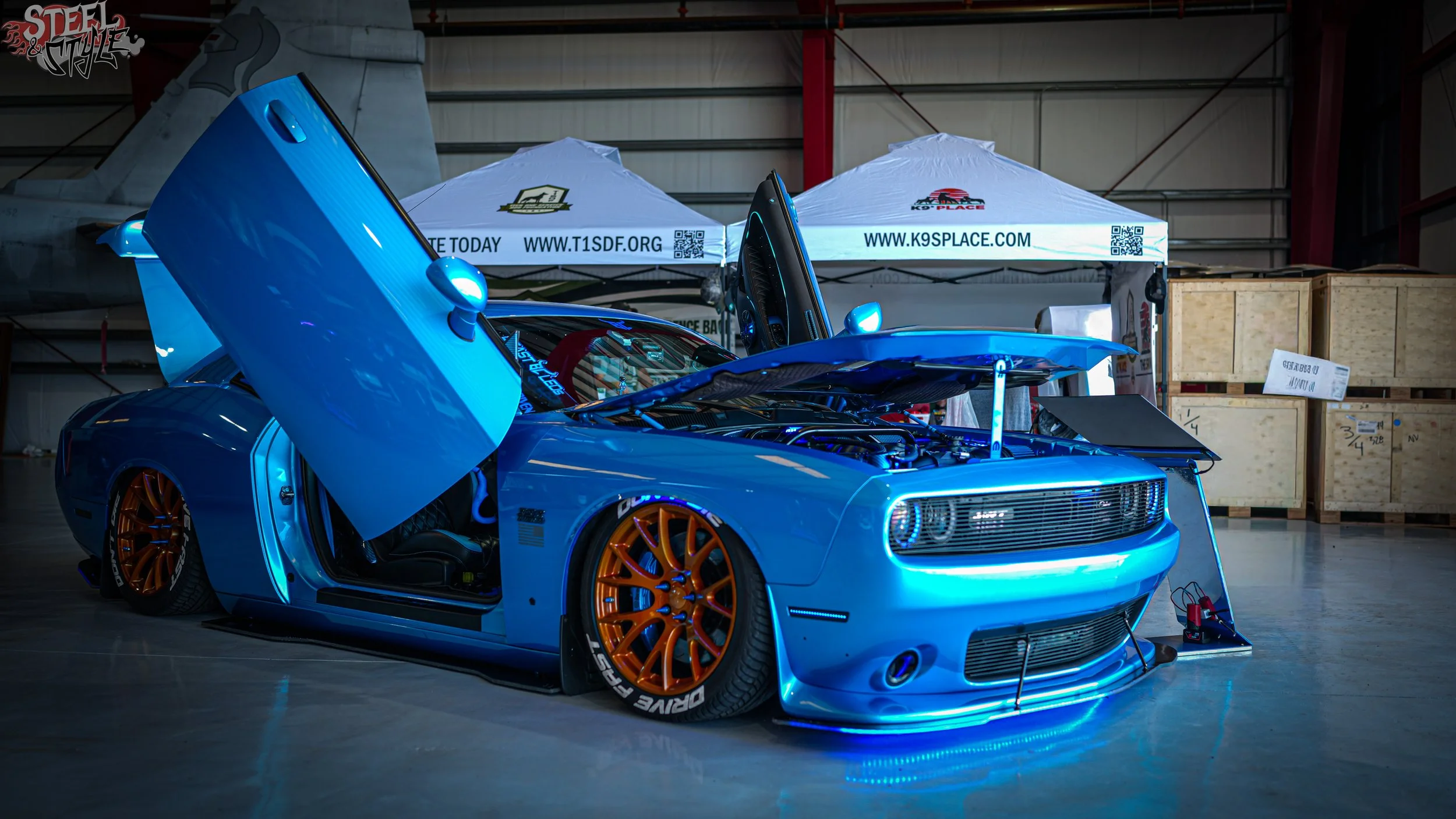 Blue sports car with open hood and doors in a garage or warehouse, with tents in the background labeled 'K9 Place' and 'T1SDF.org'.