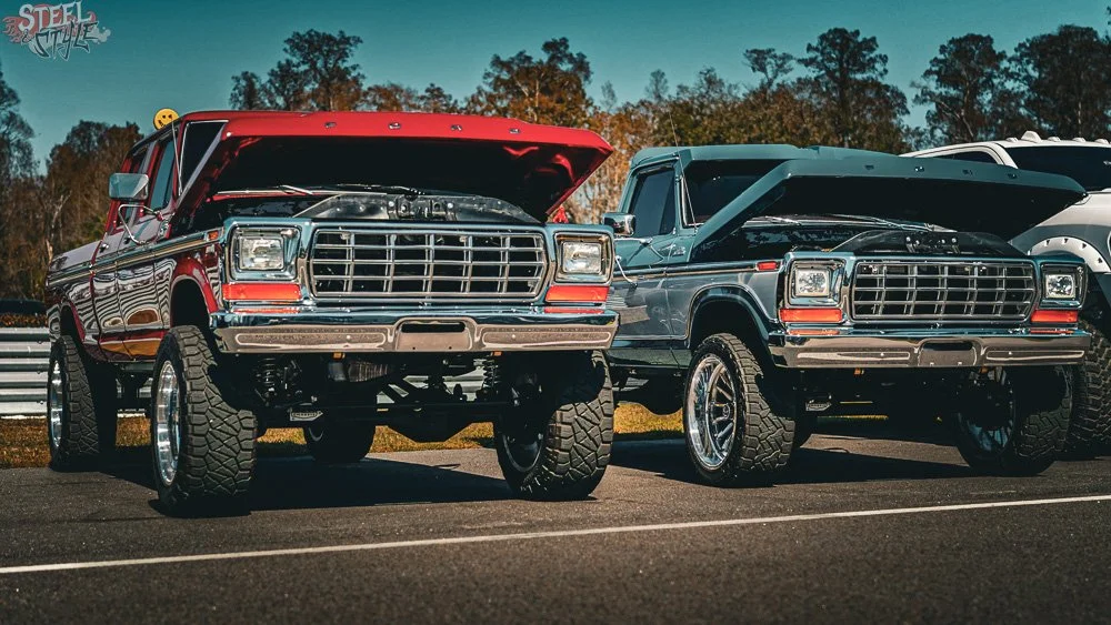 Two vintage pickup trucks with open hoods parked side by side at an outdoor car show. One is red with large off-road tires, and the other is dark green with custom rims. Trees and a partly cloudy sky are in the background.