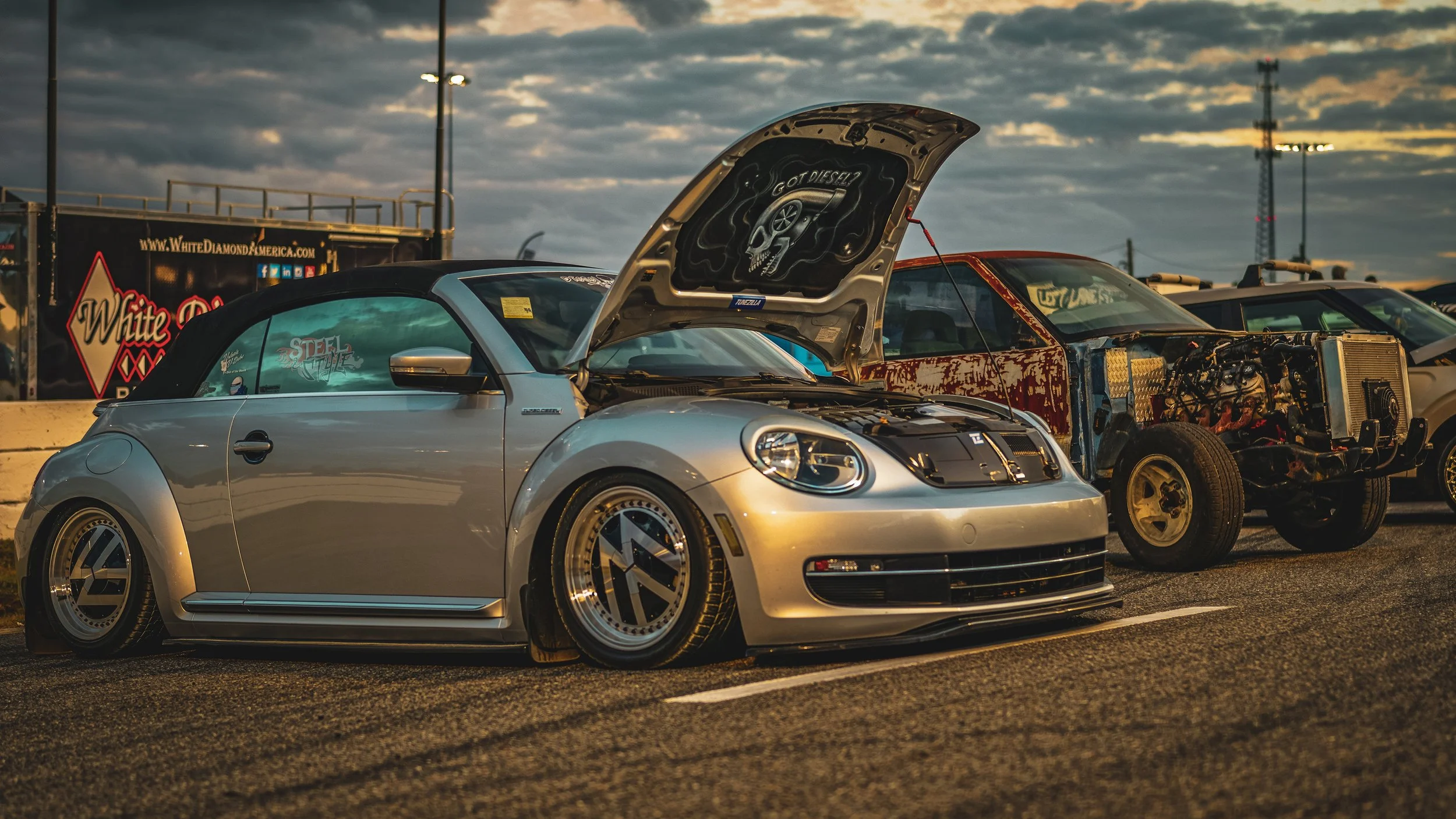 Silver lowered Volkswagen Beetle with a black convertible top parked at a race track during sunset, with other cars and a trailer in the background.