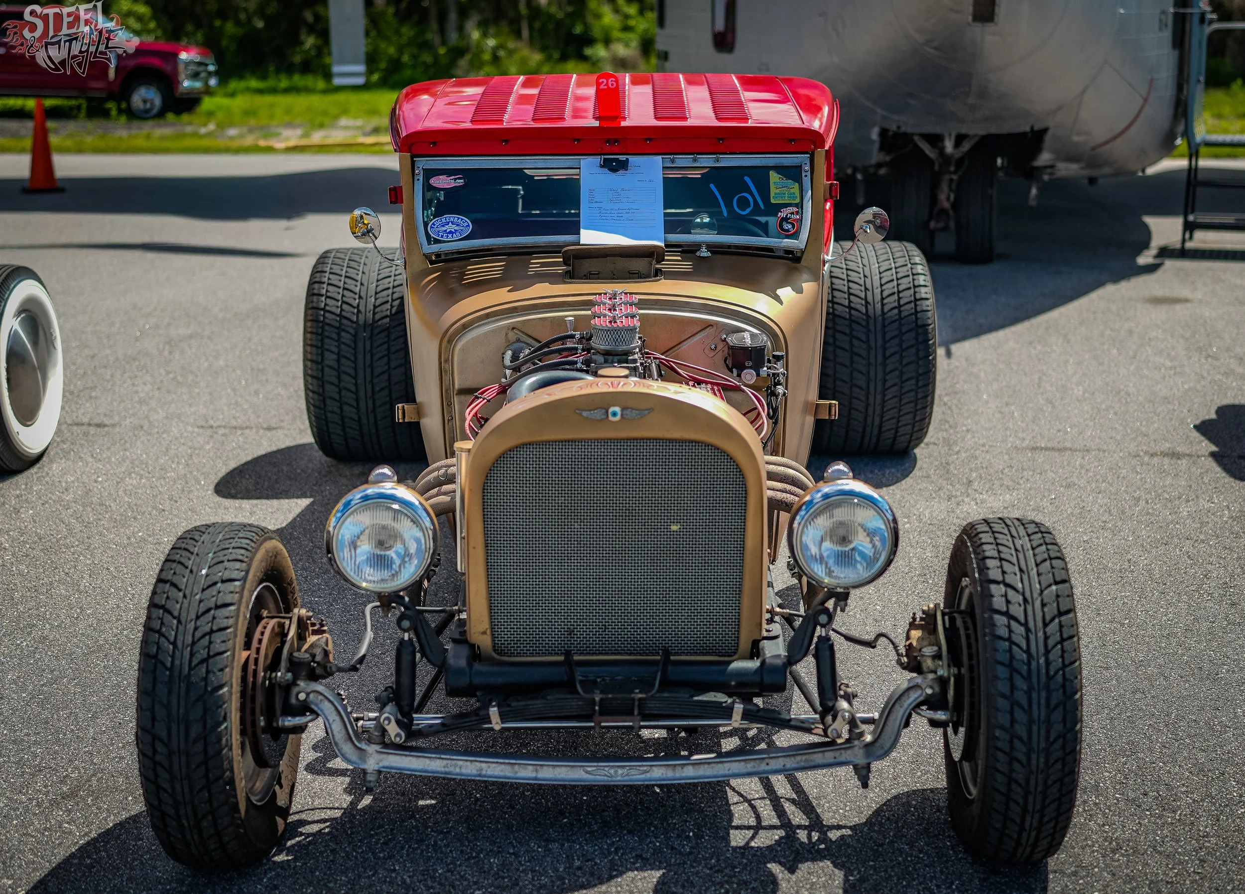 Front view of a vintage race car with exposed engine, large tires, and a red roof, parked outdoors in a parking lot with traffic cones and a trailer in the background.