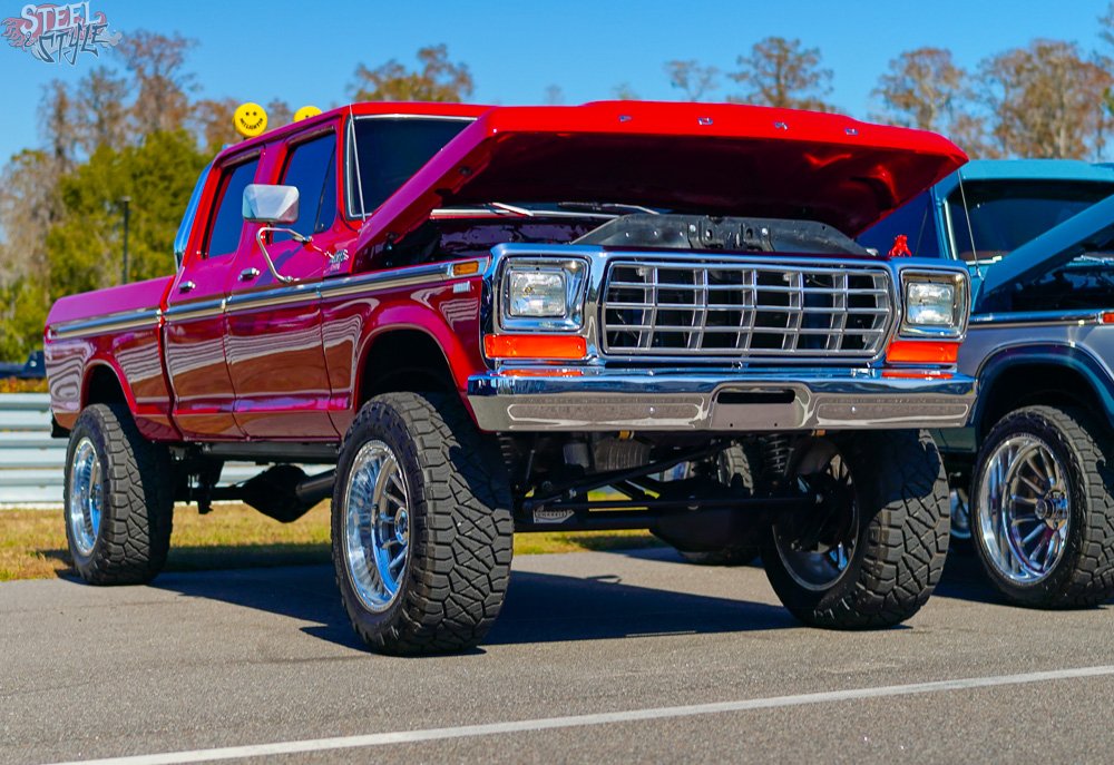 A lifted red Ford truck with its hood open parked at an outdoor car show, with other vehicles and trees in the background.