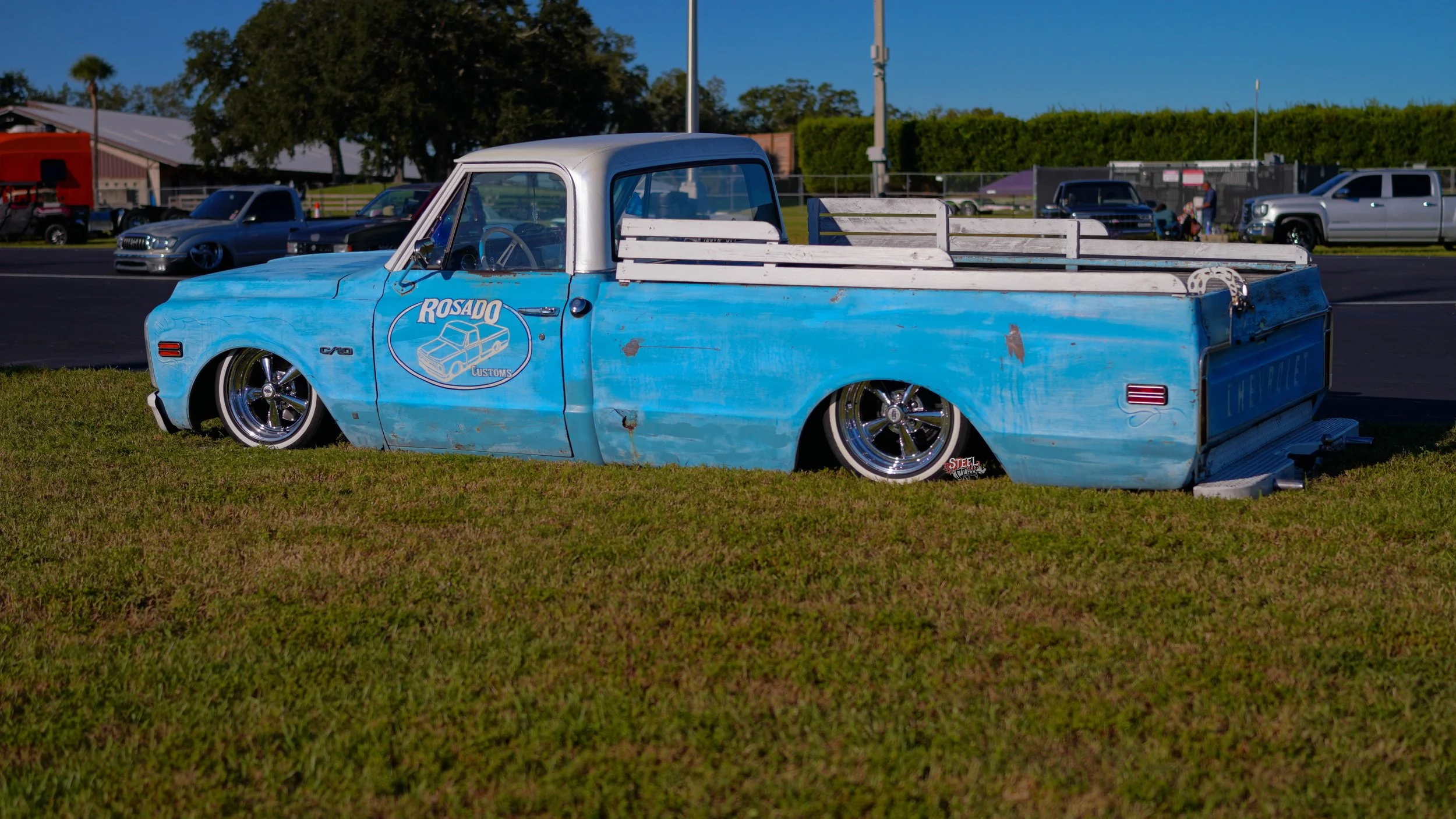 An old, weathered blue pickup truck with the logo 'Rosado Customs' on its door, parked on grass with other cars in the background, at what appears to be a car show.