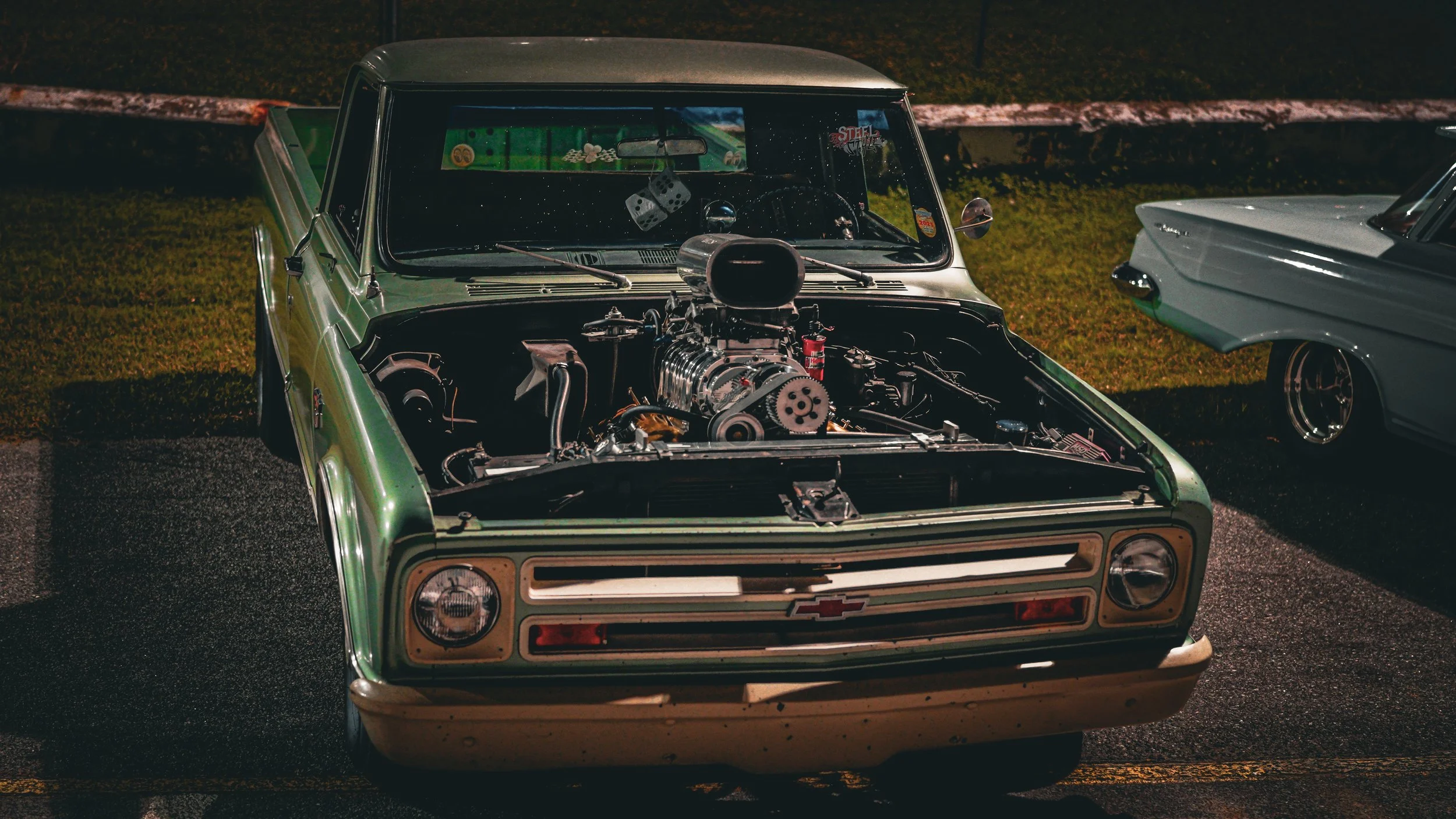 A vintage green pickup truck with an exposed engine on display at night, parked beside another classic car.