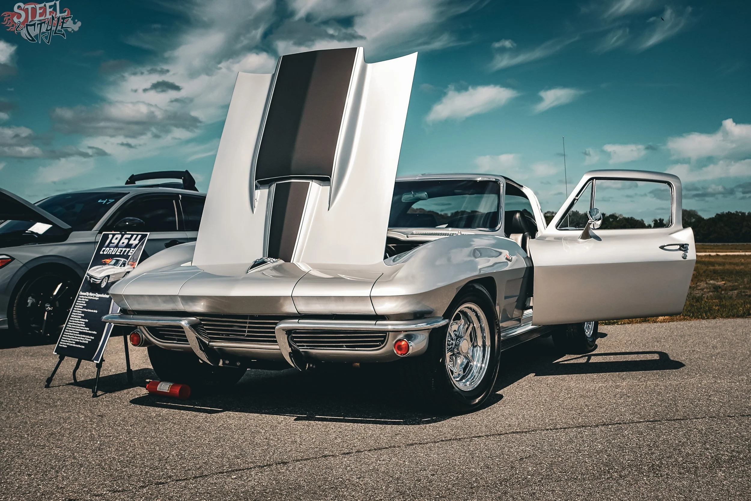 A classic silver Corvette with its hood and driver side door open at an outdoor car show, with a information stand and a sign in front, under a partly cloudy sky.