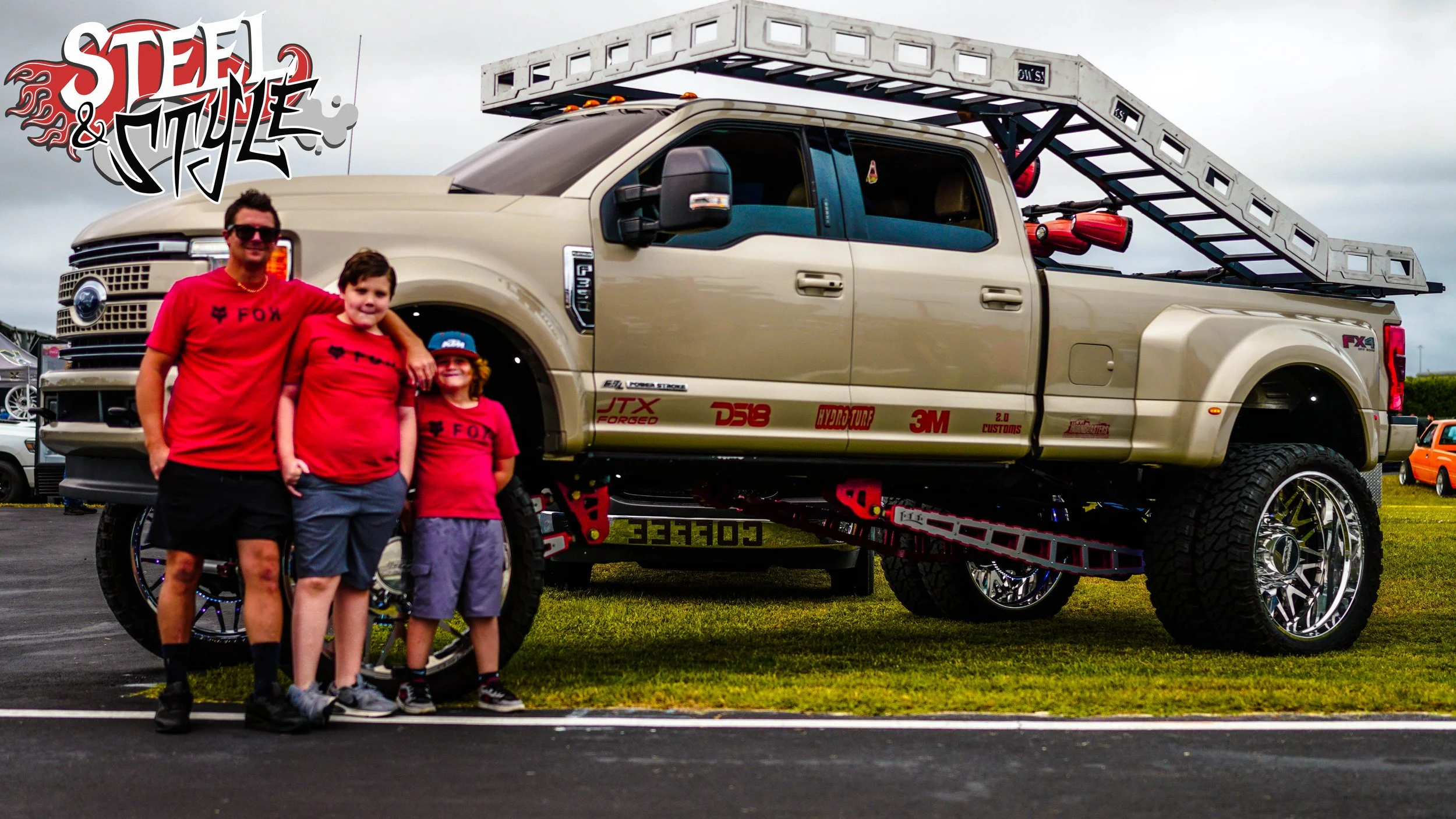 Three children and one adult standing in front of a large modified pickup truck with a ladder on its bed. The children are wearing red shirts with the FOX logo, and the adult is wearing a red shirt. The truck has big wheels, decals, and custom parts,