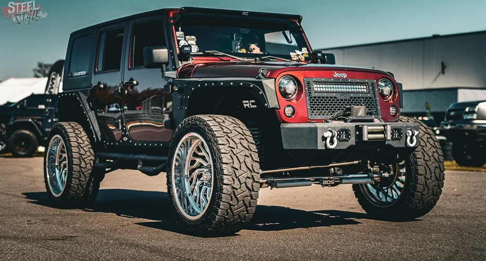 Customized Jeep with large off-road tires, black and red paint, and aftermarket front bumper, parked outdoors in a lot.