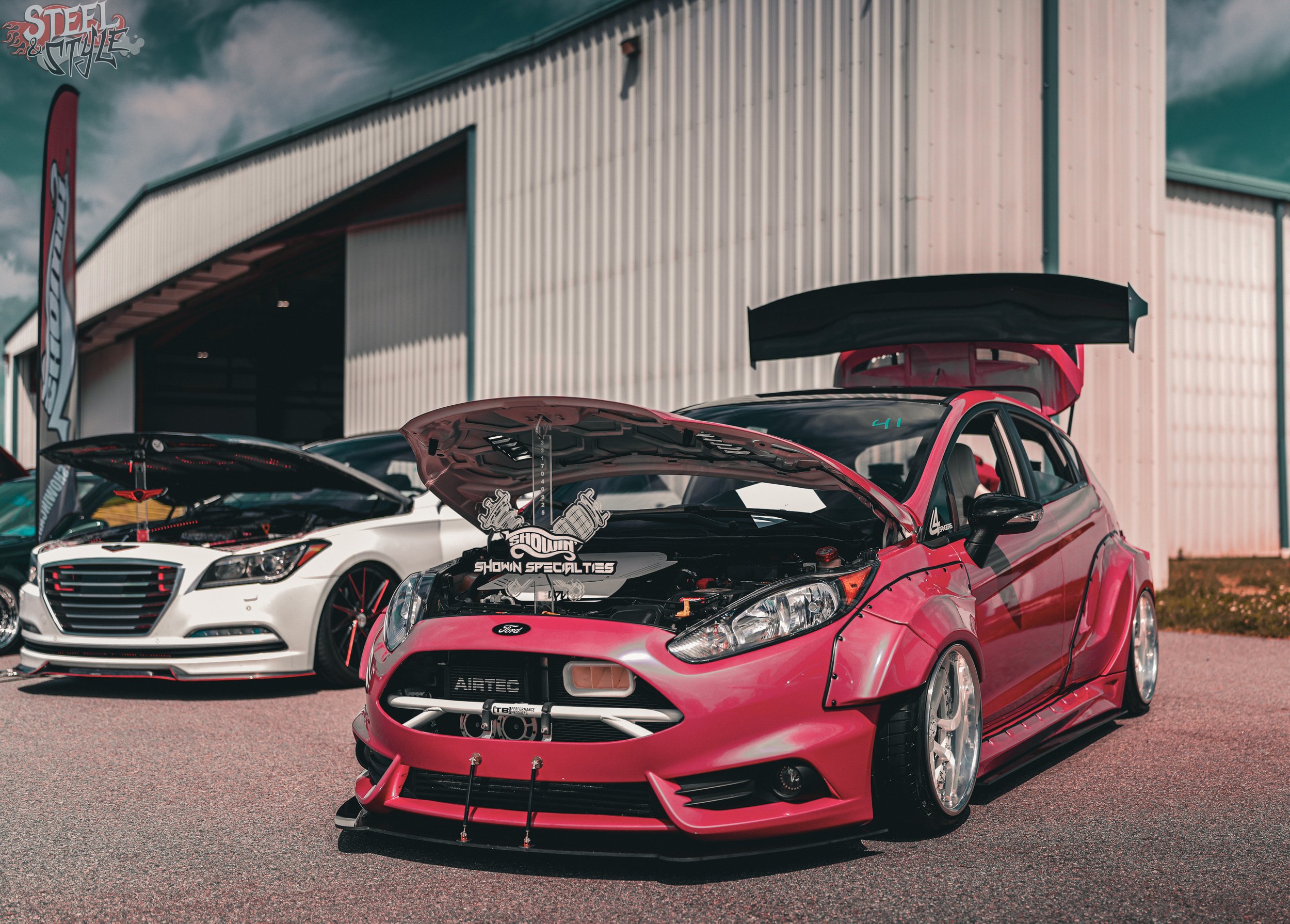 Red and black sports car with open hood and large rear wing on display at a car show, with other vehicles and a building with corrugated metal siding in the background.