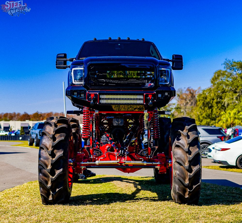 A monster truck with large tires, a black cab, and red suspension parts parked on grass during daytime.