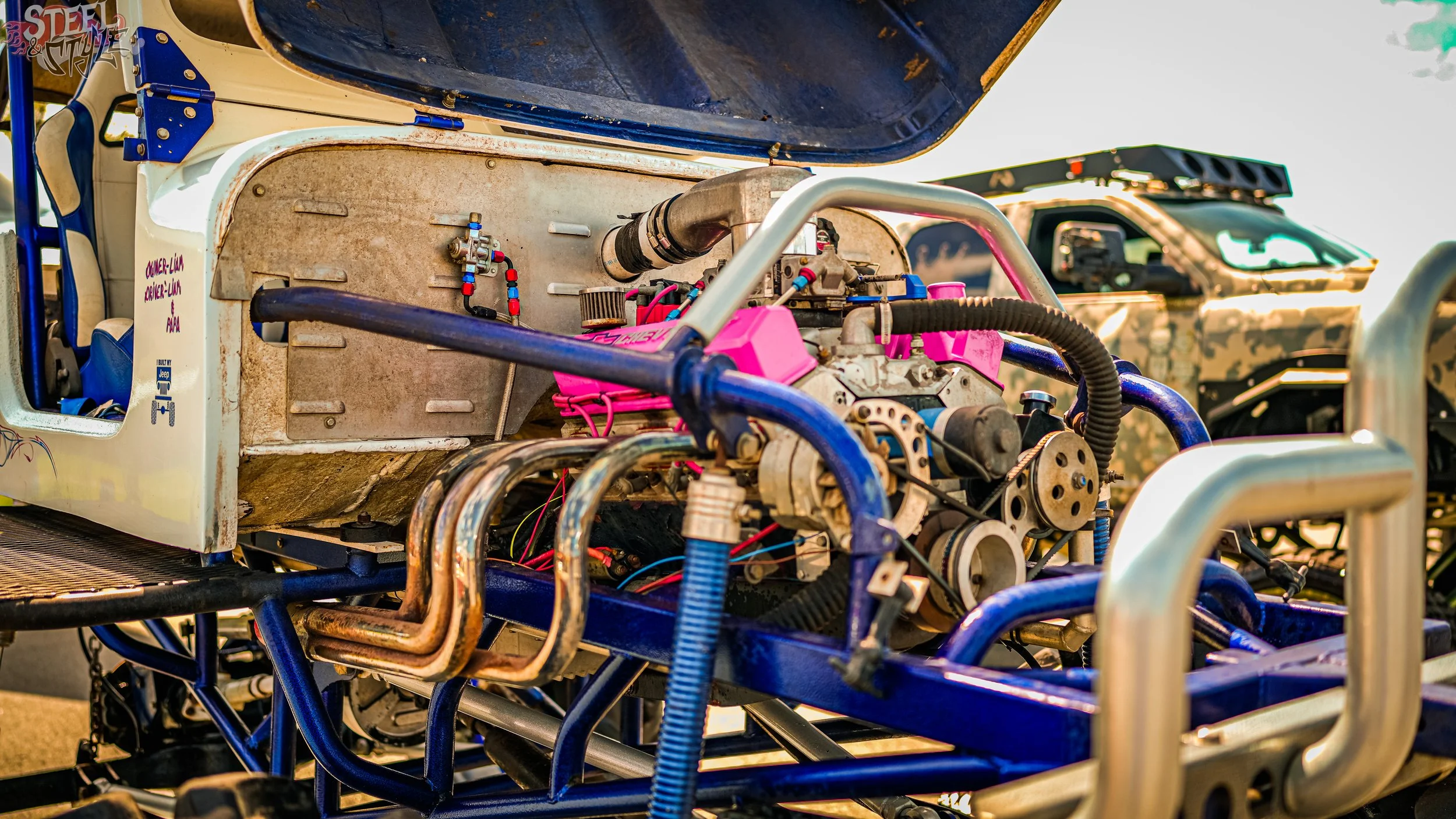 Close-up of a race car's engine with exposed parts and wiring, showing the engine bay and surrounding frame, with a truck in the background.